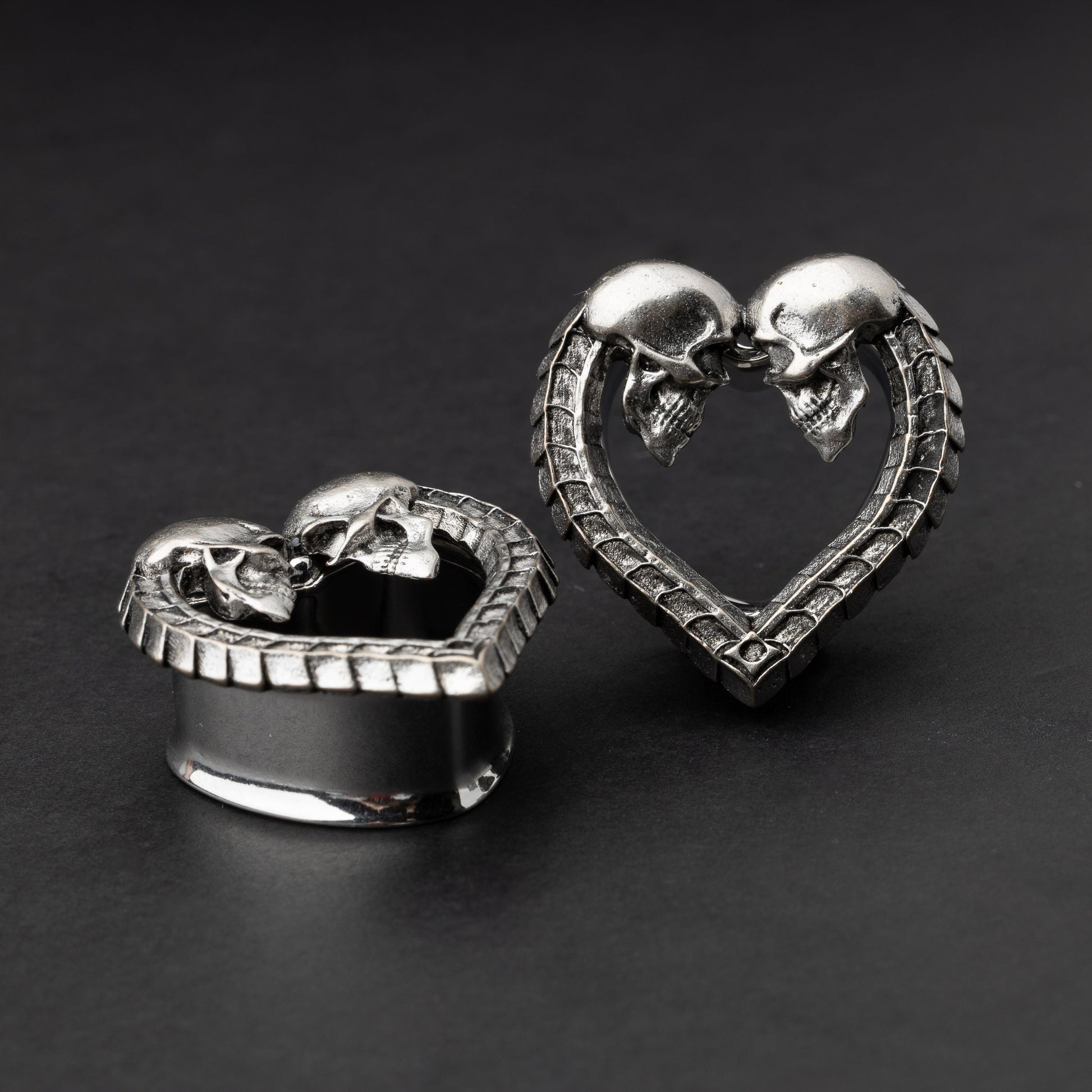 Heart-shaped tunnel plugs with intricate skull details and textured edges, displayed on a dark background.