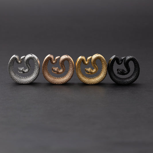 Snake-shaped saddle plugs in stainless steel, gold, rose gold, and black finishes, displayed on a dark background.