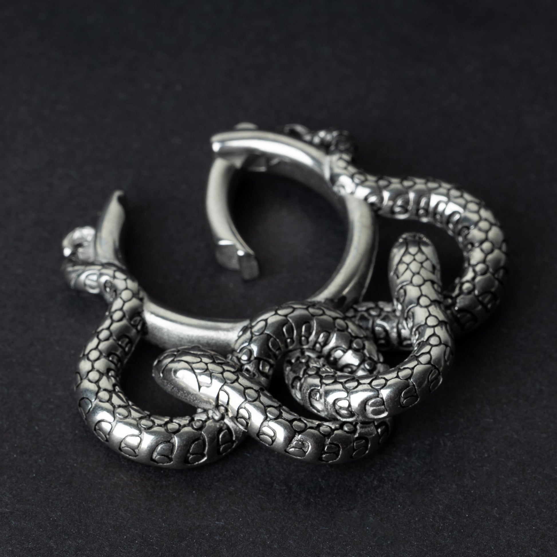 One open snake ear weight in polished silver, featuring detailed scales and a seamless hinge closure, displayed on a dark background.
