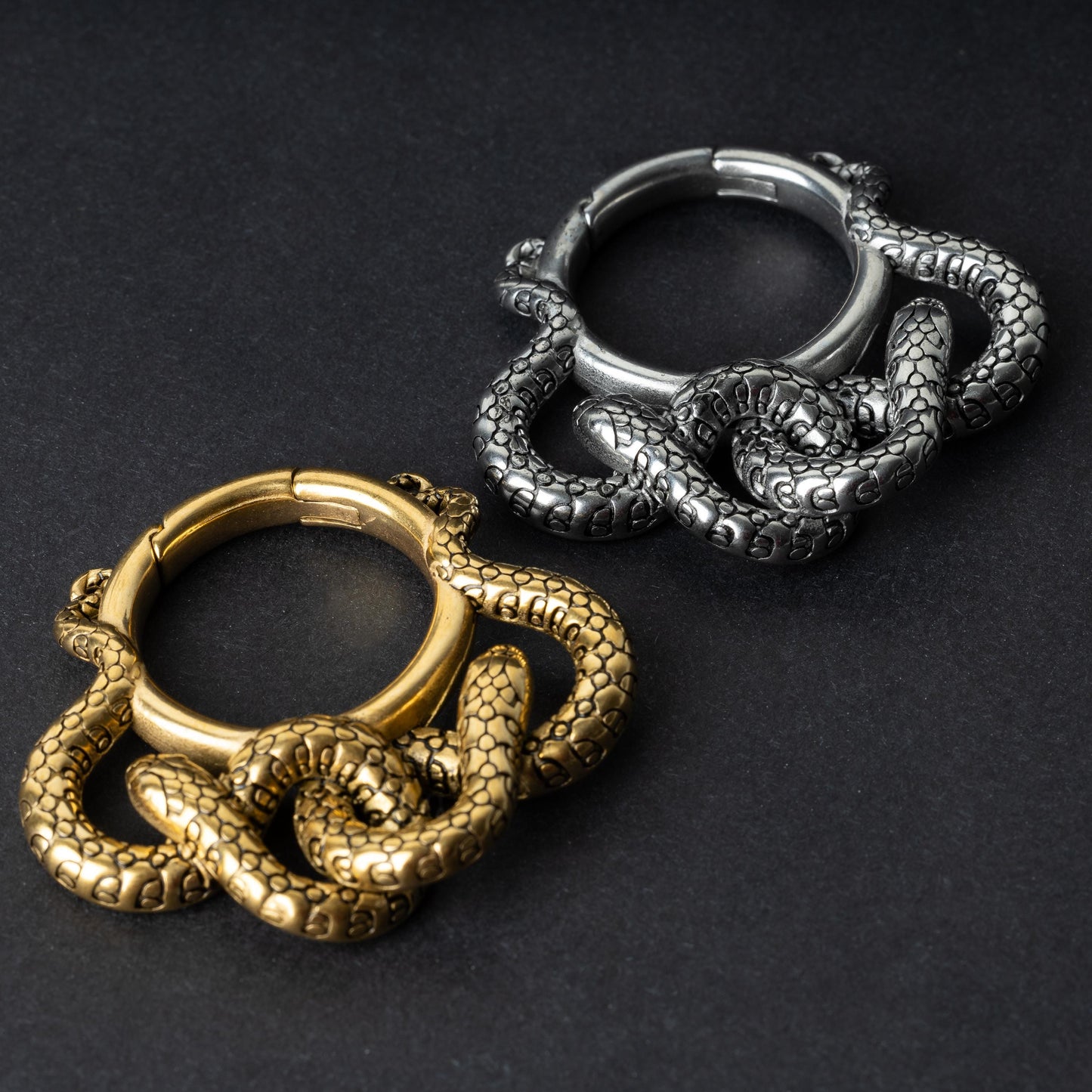 Two snake ear weights, one in polished gold and one in polished silver, featuring detailed scales and a seamless hinge closure, displayed on a dark background.

