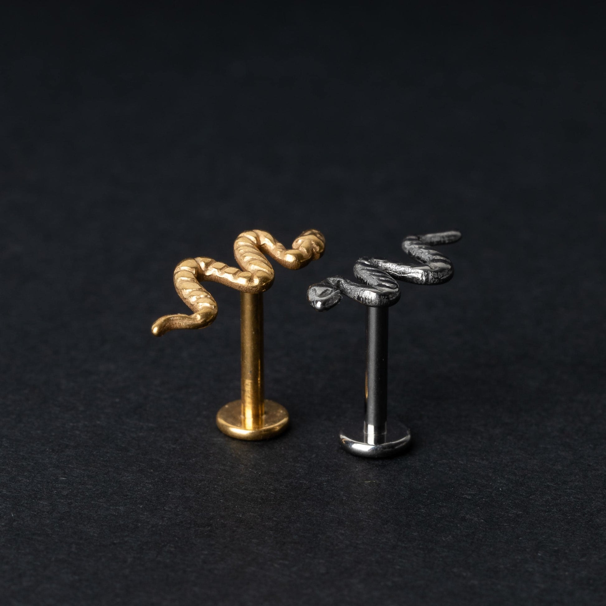 Gold-tone and silver-tone snake labret bars with intricate coiled snake designs displayed on a dark background.

