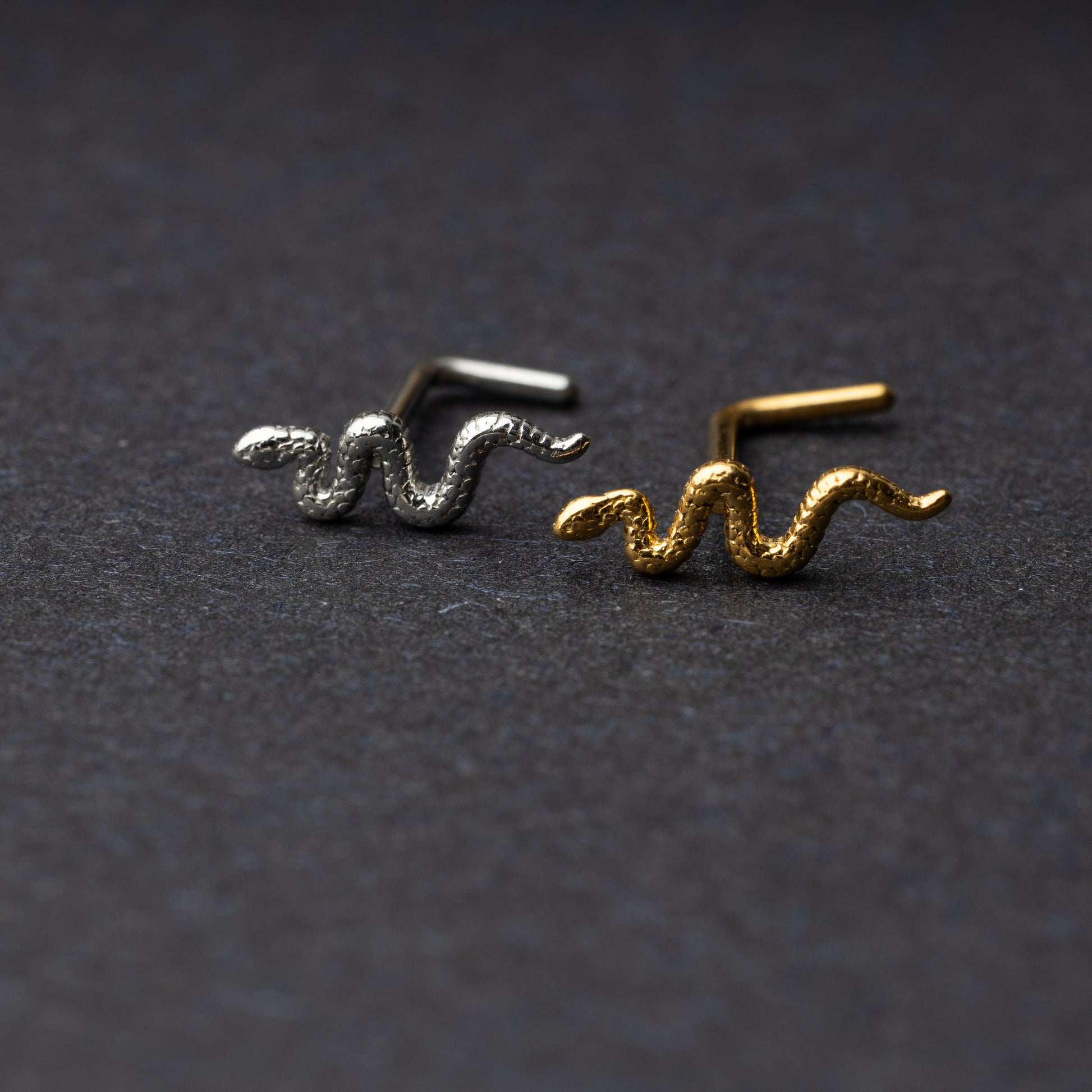 Snake-shaped nose studs in gold and silver finishes displayed on a dark background.