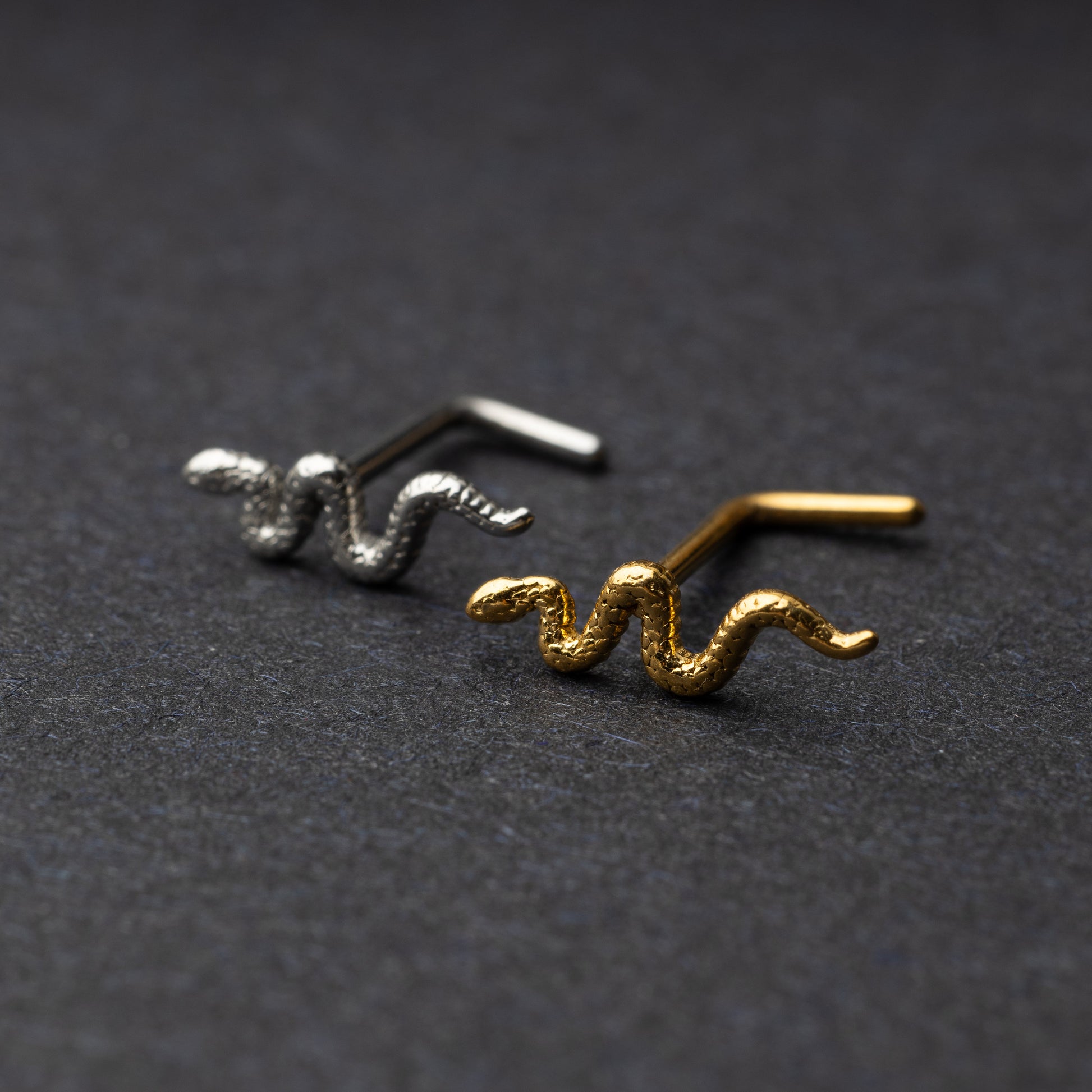 Snake-shaped nose studs in gold and silver finishes displayed on a dark background.