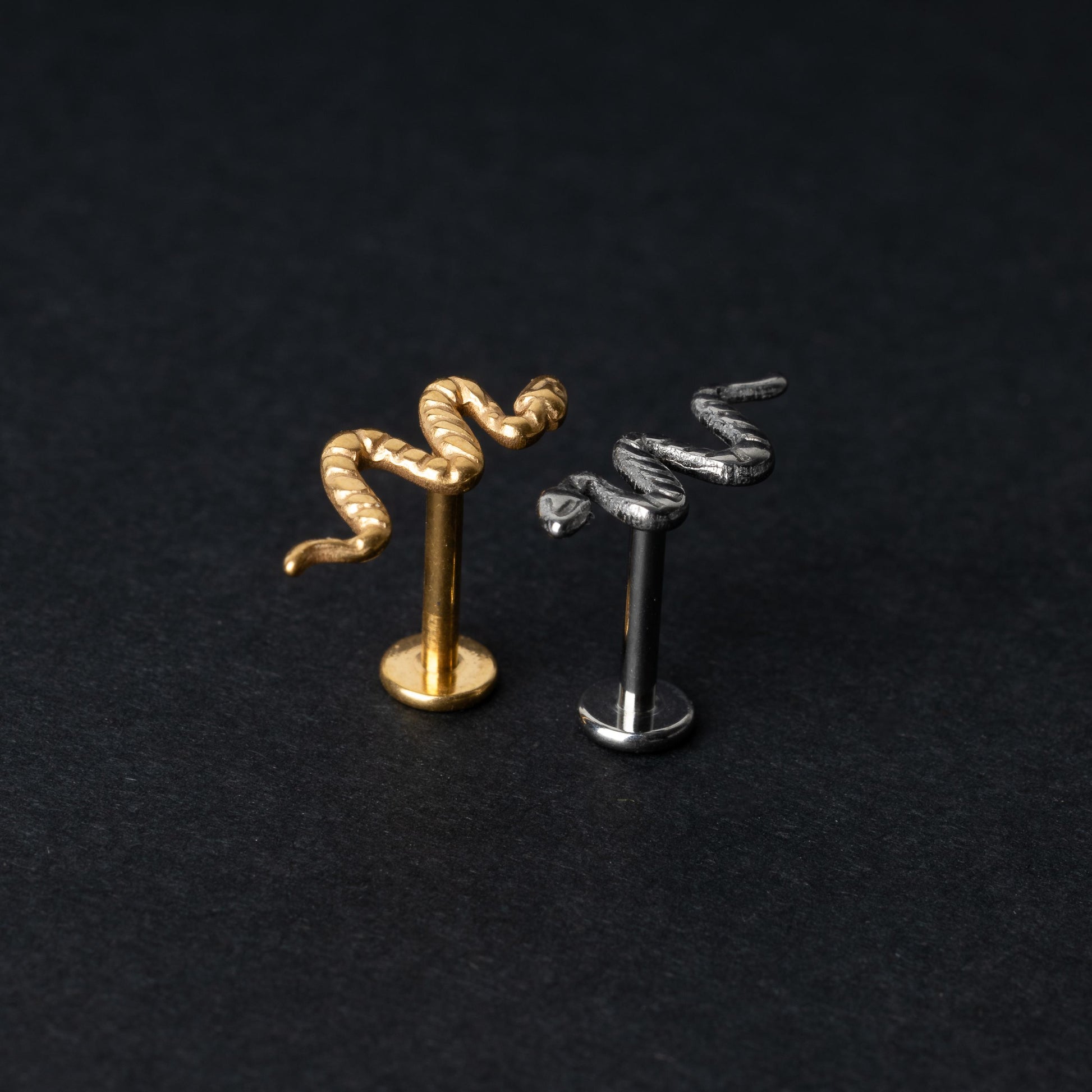 Gold-tone and silver-tone snake labret bars with intricate coiled snake designs displayed on a dark background.

