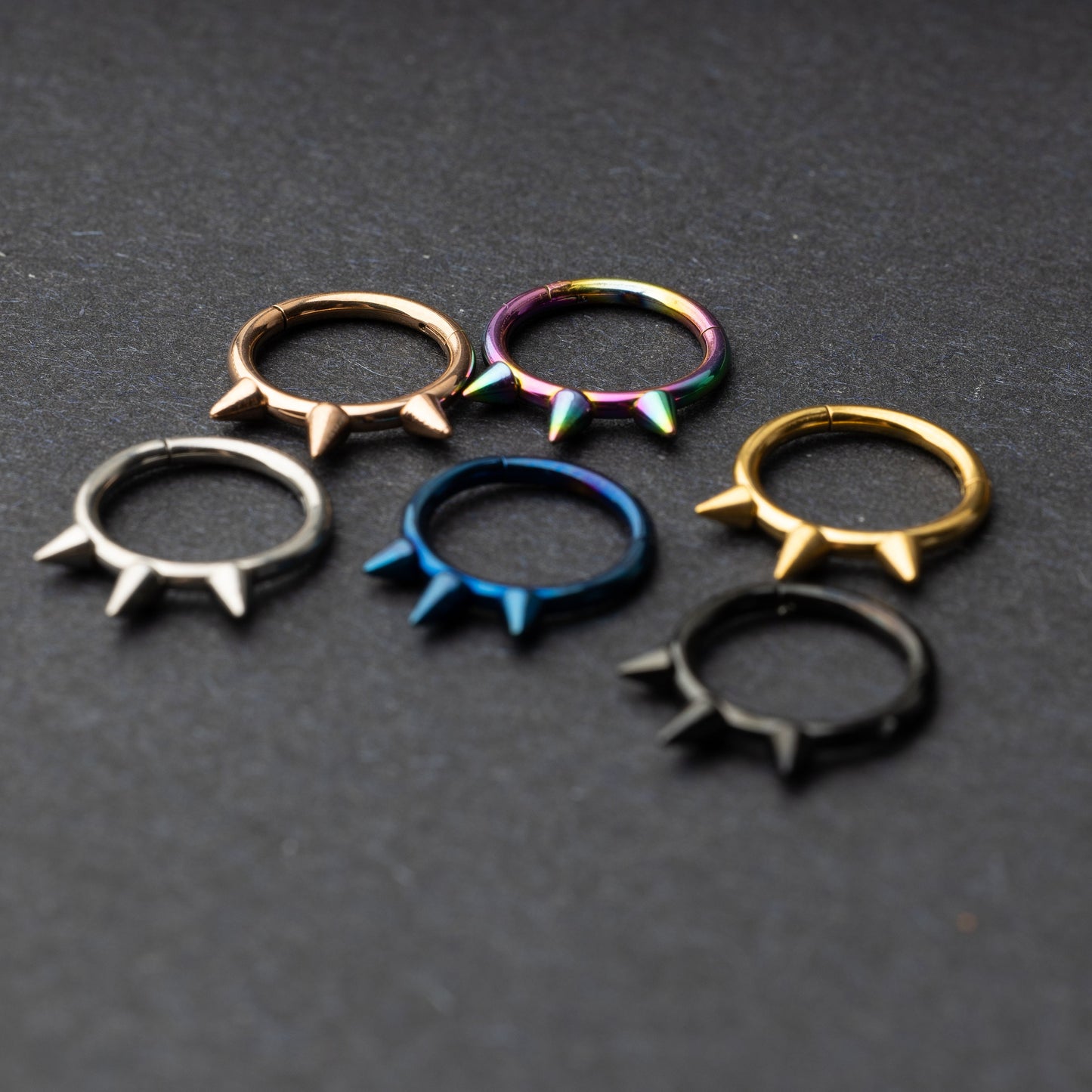 Assorted stainless steel spiked rings in various finishes, including gold, black, rainbow, and blue, displayed on a dark textured background.