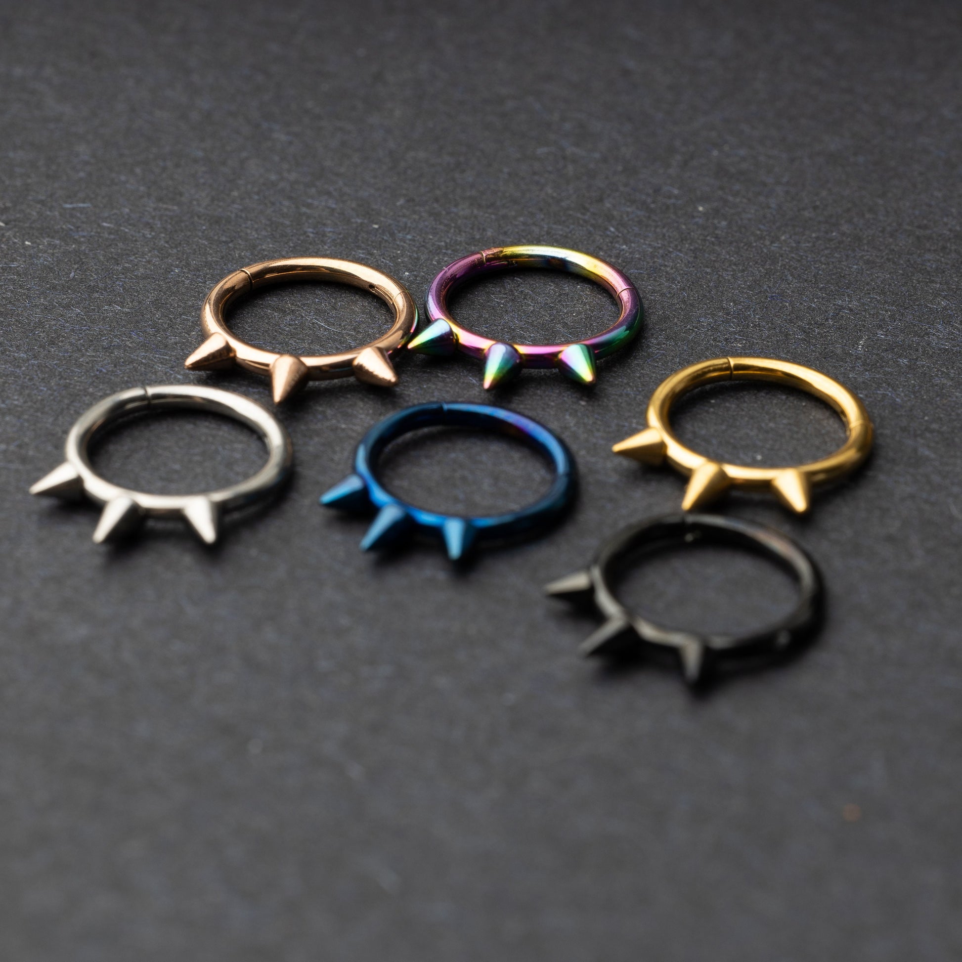 Assorted stainless steel spiked rings in various finishes, including gold, black, rainbow, and blue, displayed on a dark textured background.