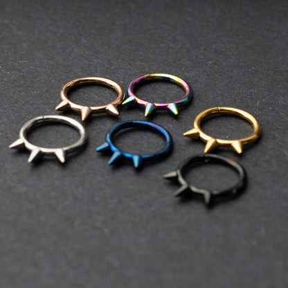 Assorted stainless steel spiked rings in various finishes, including gold, black, rainbow, and blue, displayed on a dark textured background.