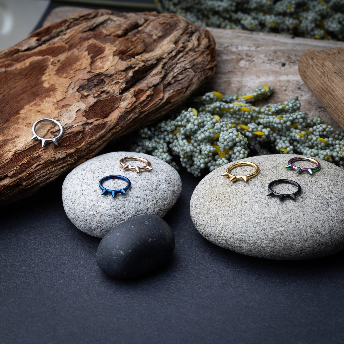 Assorted stainless steel spiked rings in various finishes, including gold, black, rainbow, and blue, displayed on a dark textured background.