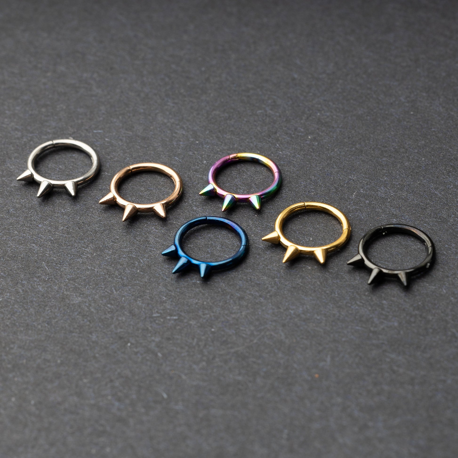 Assorted stainless steel spiked rings in various finishes, including gold, black, rainbow, and blue, displayed on a dark textured background.