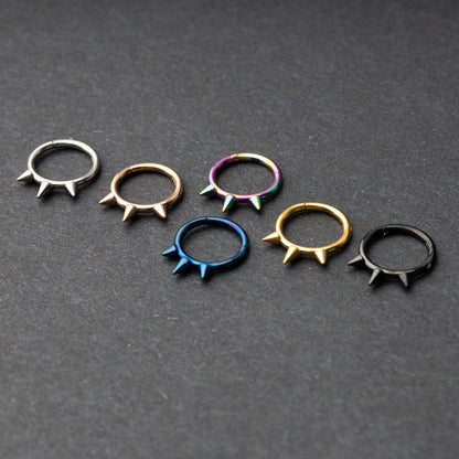 Assorted stainless steel spiked rings in various finishes, including gold, black, rainbow, and blue, displayed on a dark textured background.