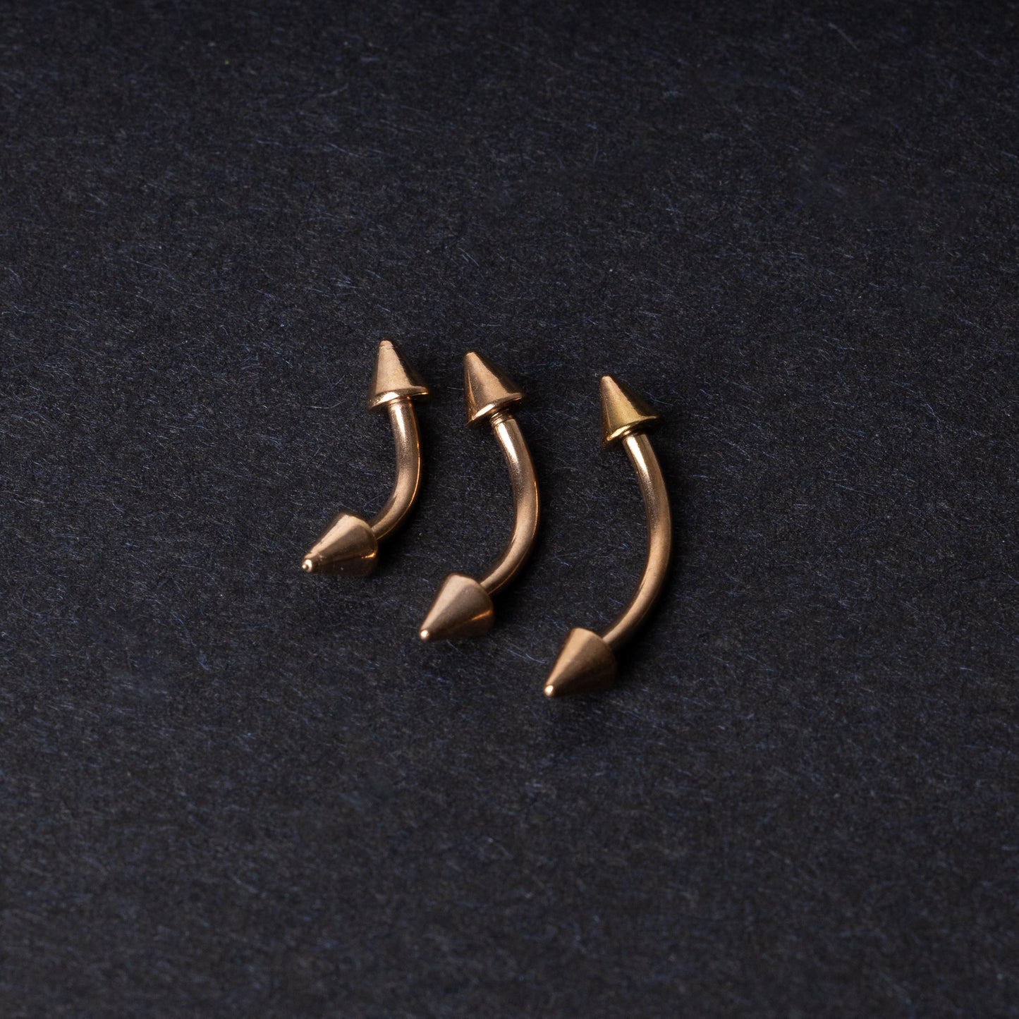 Three gold-tone titanium curved barbells with cone ends, displayed on a dark textured background, showcasing their sleek curved design and polished finish.