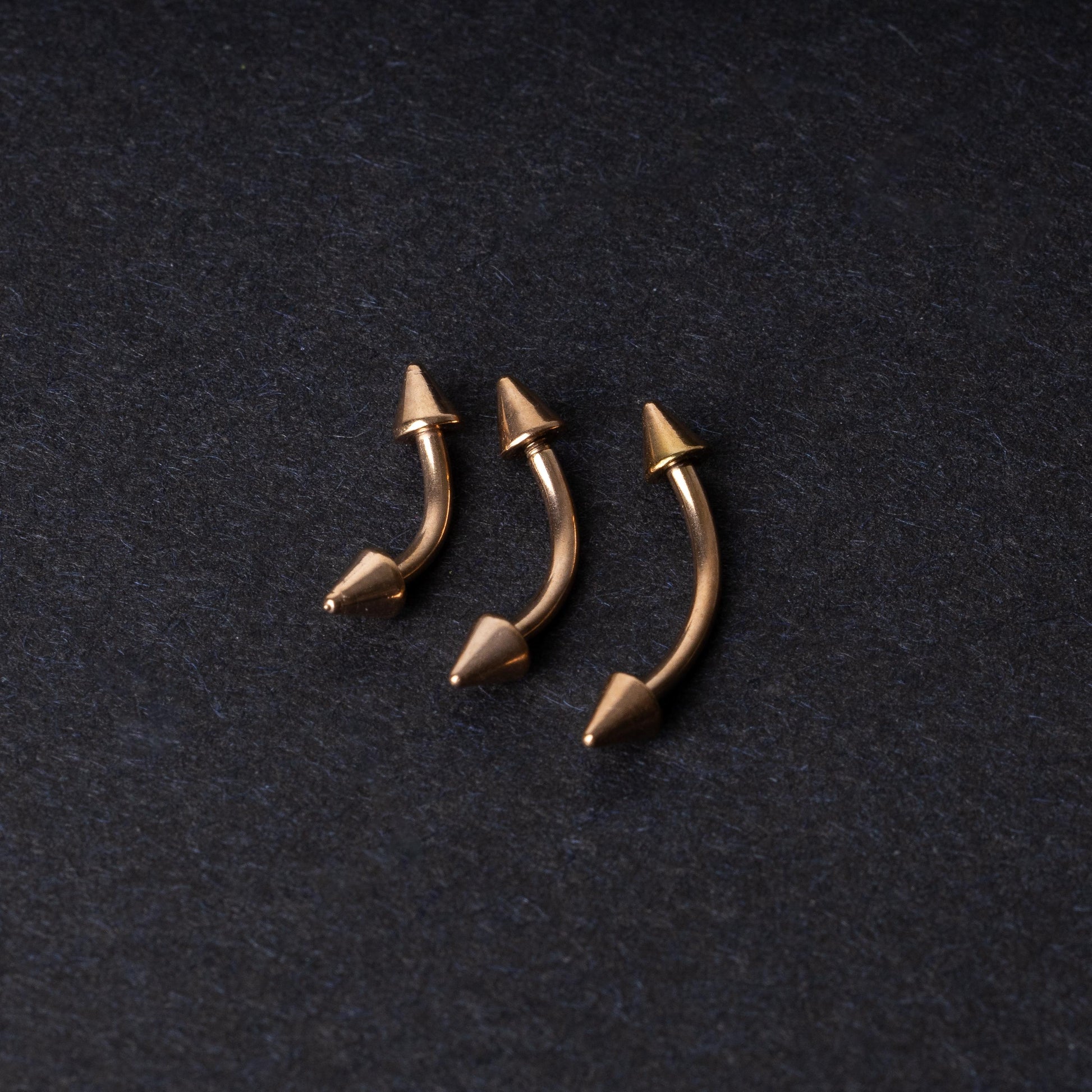 Three gold-tone titanium curved barbells with cone ends, displayed on a dark textured background, showcasing their sleek curved design and polished finish.