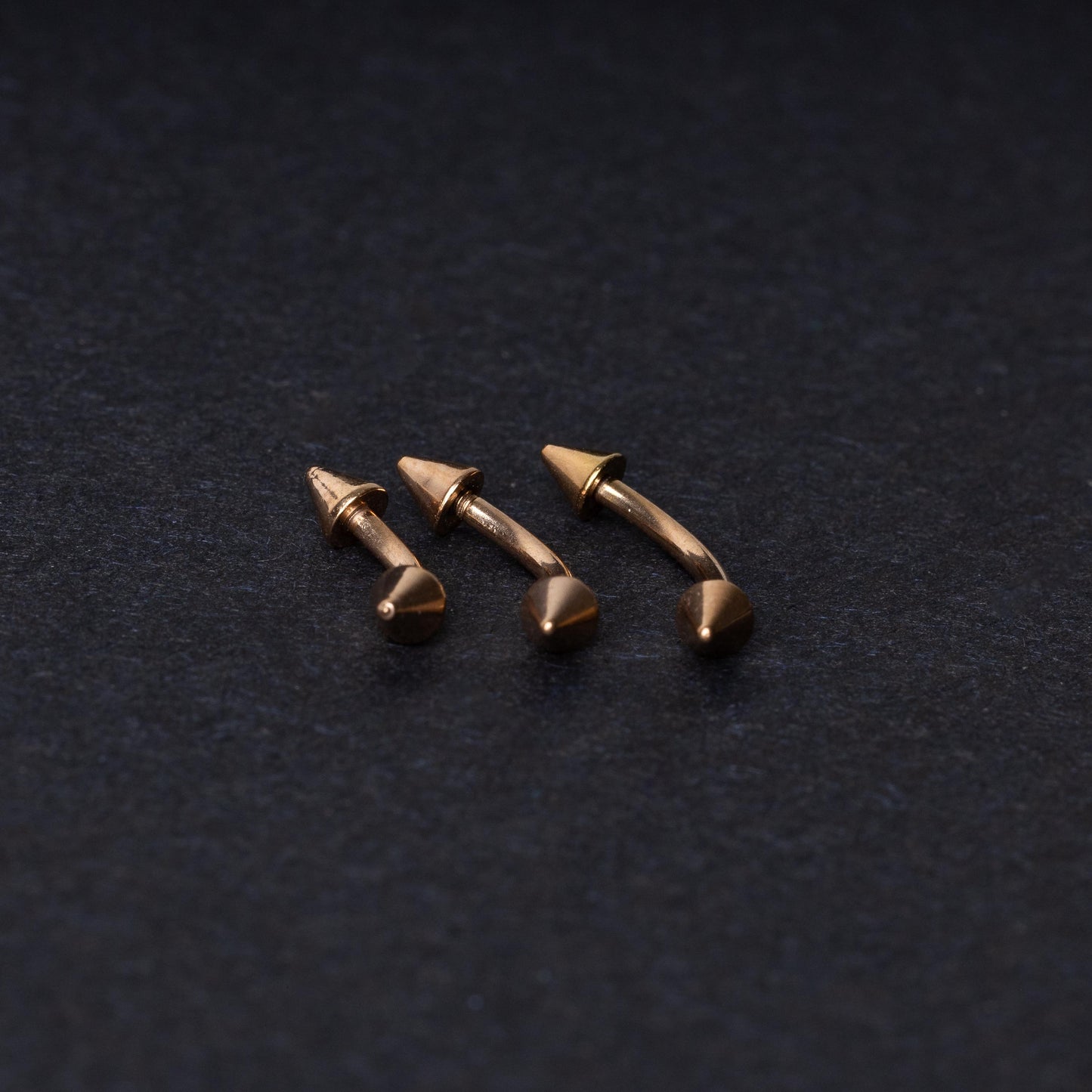 Three gold-tone titanium curved barbells with cone ends, displayed on a dark textured background, showcasing their sleek curved design and polished finish.