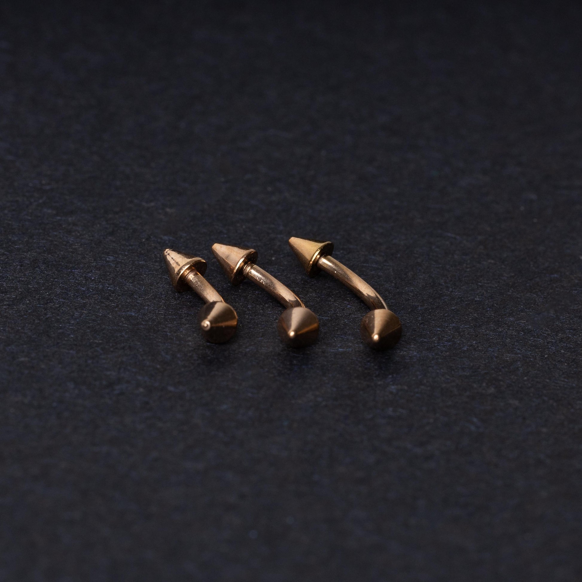 Three gold-tone titanium curved barbells with cone ends, displayed on a dark textured background, showcasing their sleek curved design and polished finish.