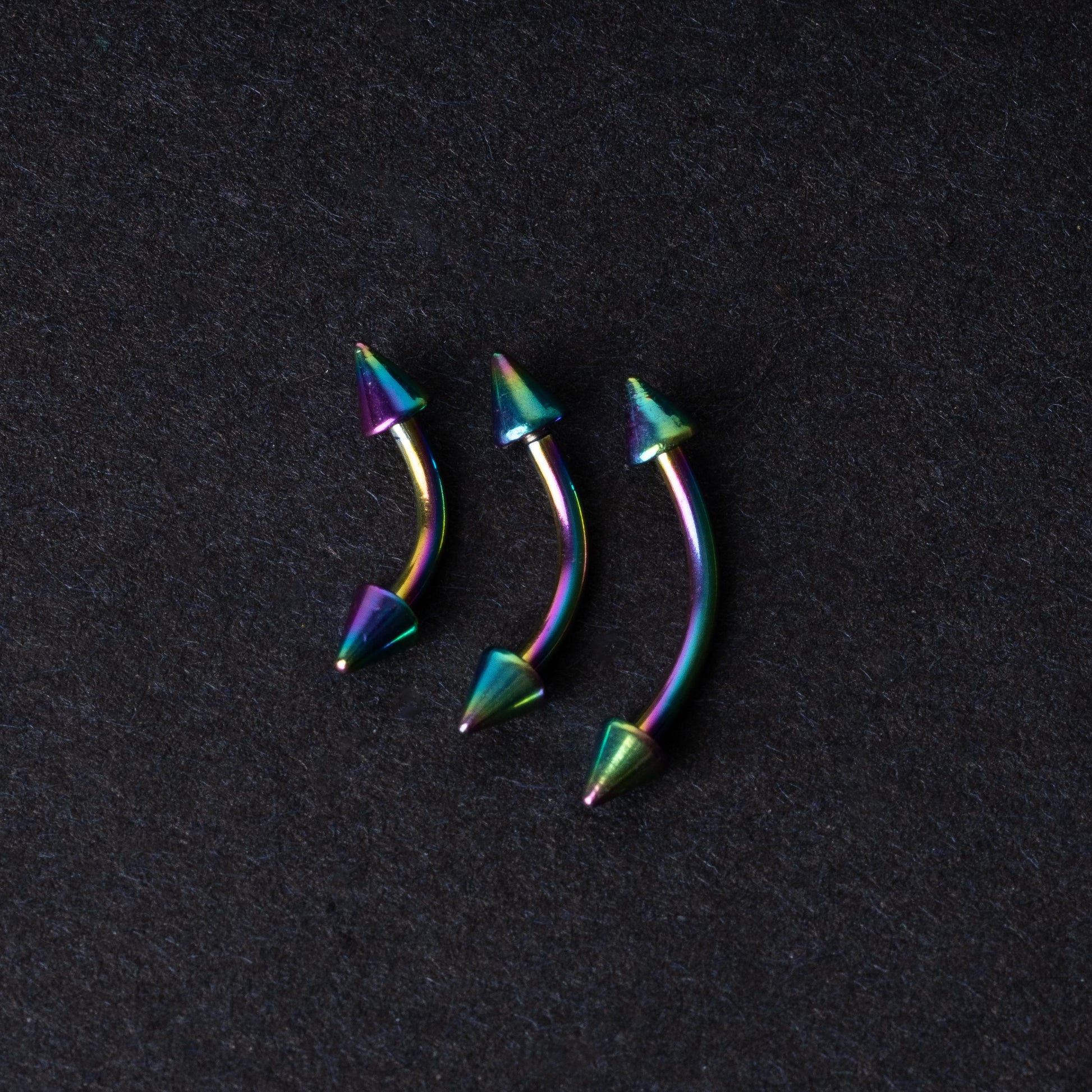 Three rainbow titanium curved barbells with cone-shaped ends, displayed on a dark textured background, showcasing their vivid color gradient and sleek design.