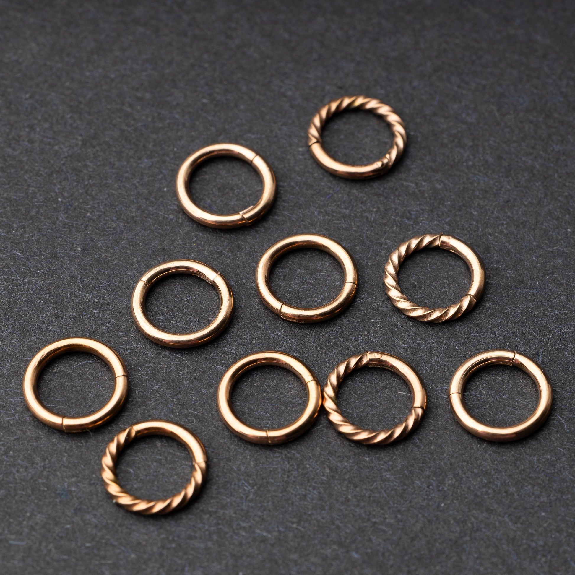 Set of rose gold clicker rings with smooth and twisted designs, displayed on a dark background.