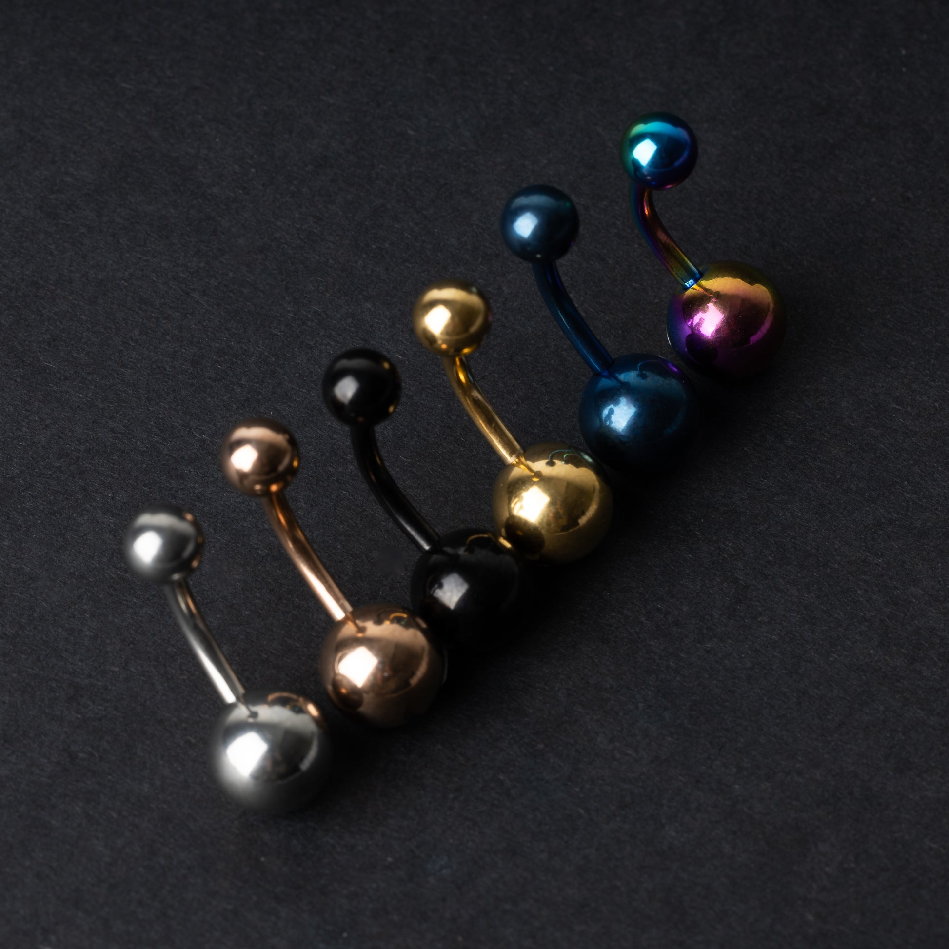 Stainless steel belly rings in silver, rose gold, black, gold, blue, and rainbow finishes, displayed on a dark background.