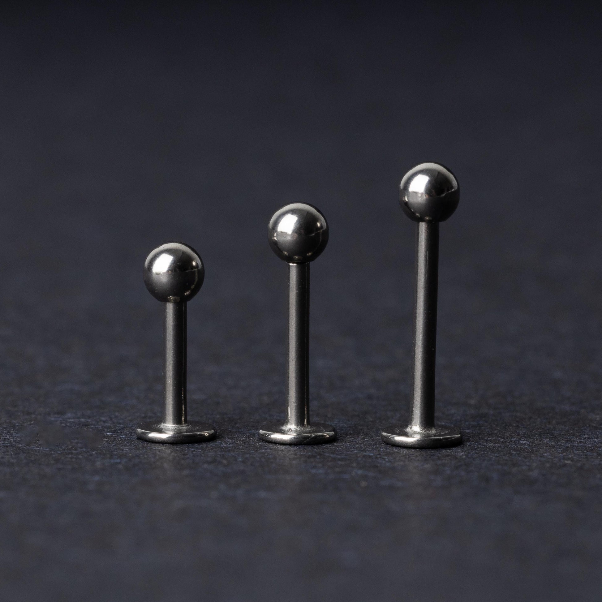 Three silver stainless steel labret studs with flat backs and polished ball ends, displayed on a dark textured background.