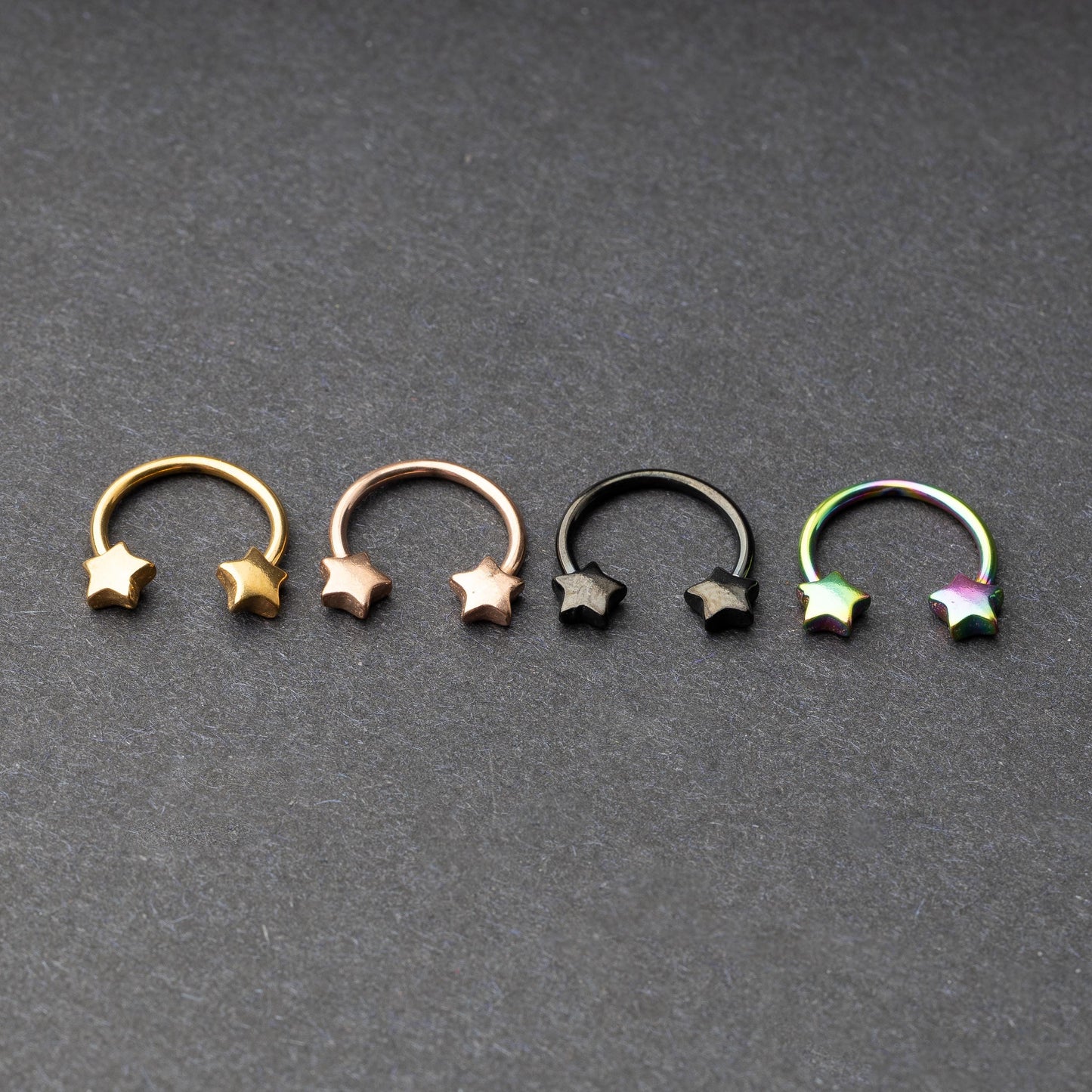 Four stainless steel horseshoe hoops with star-shaped ends in gold, rose gold, black, and rainbow finishes displayed on a dark background.