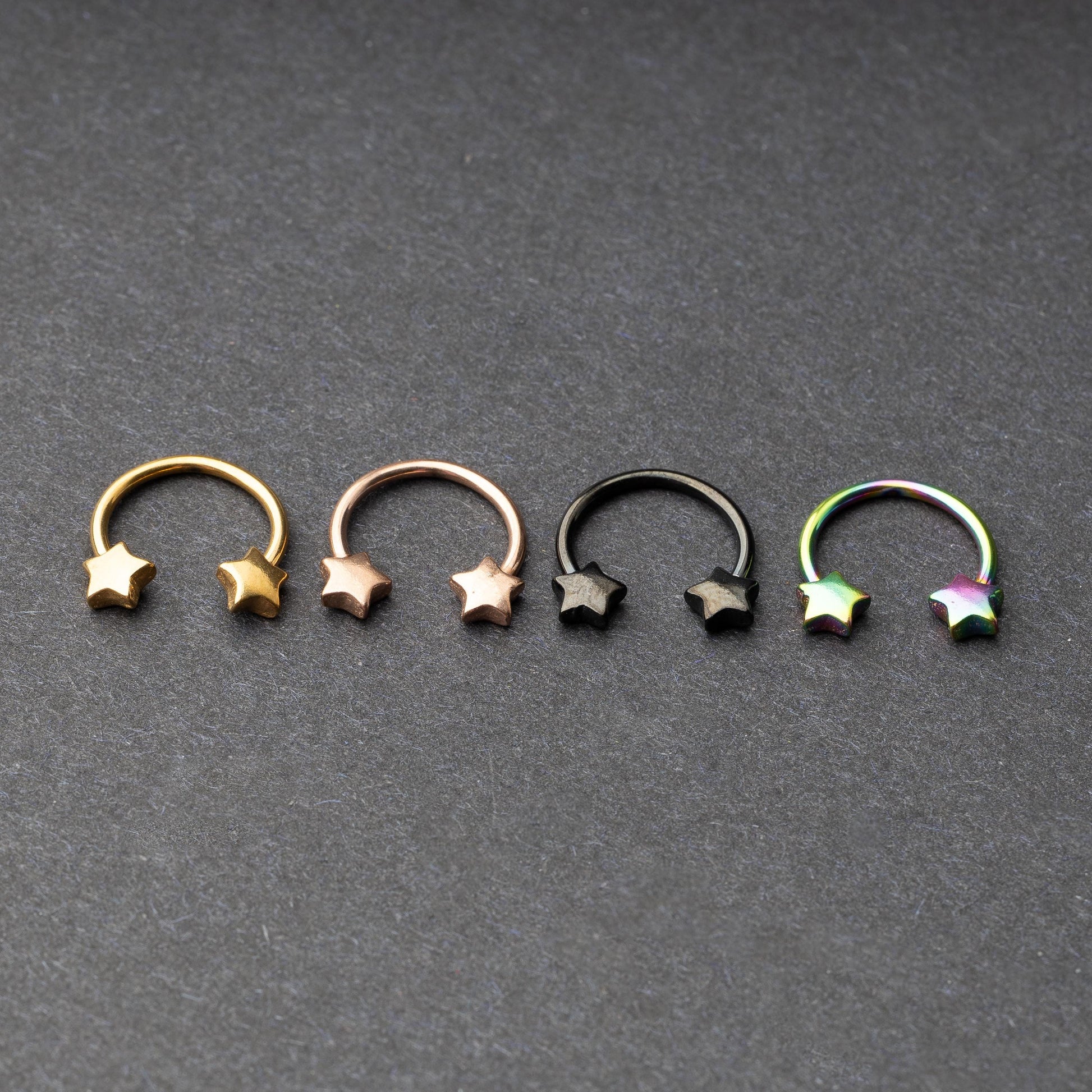 Four stainless steel horseshoe hoops with star-shaped ends in gold, rose gold, black, and rainbow finishes displayed on a dark background.