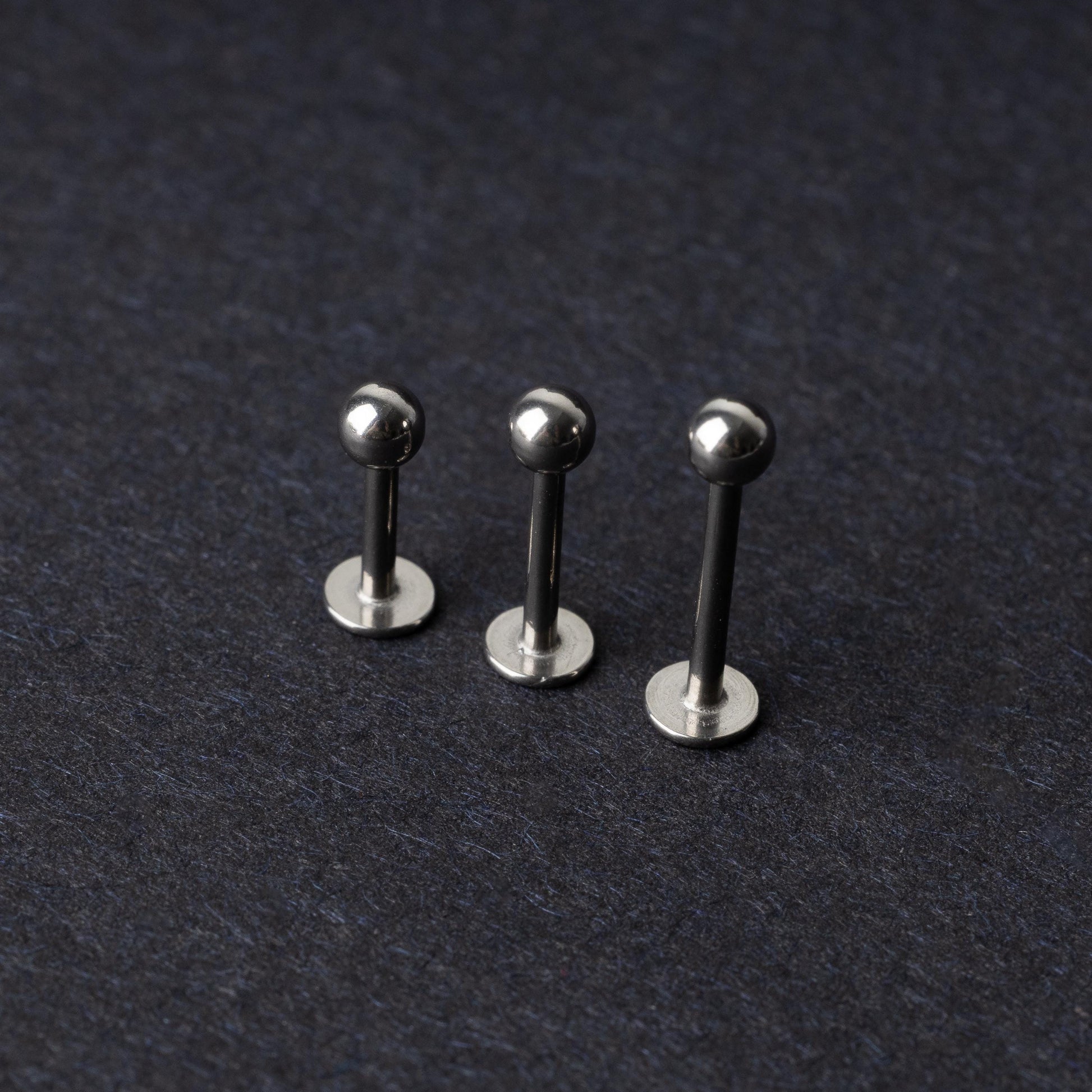Three silver stainless steel labret studs with flat backs and polished ball ends, displayed on a dark textured background.