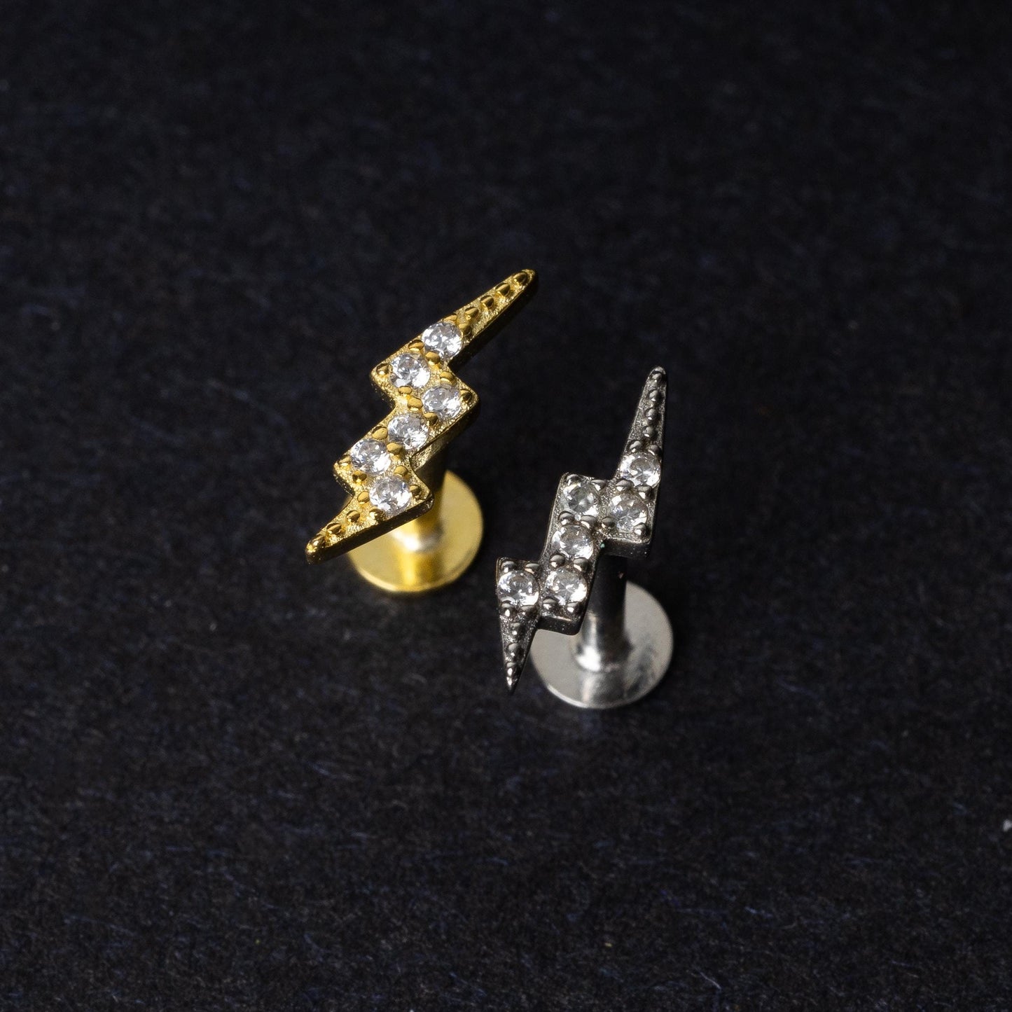 Gold and silver lightning bolt labret studs with crystal detailing, displayed on a dark background.