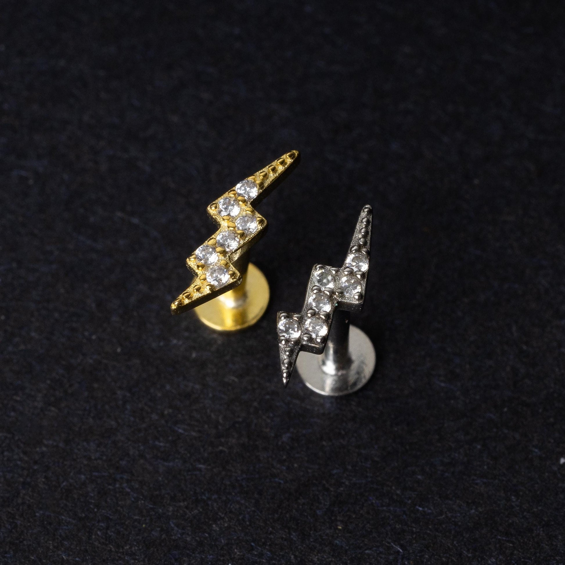 Gold and silver lightning bolt labret studs with crystal detailing, displayed on a dark background.