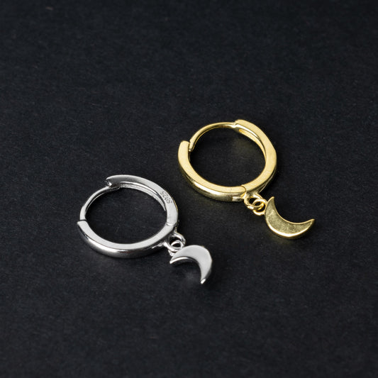 Two crescent moon huggie hoop earrings in gold and silver, each featuring a dangling crescent charm, displayed on a dark background.