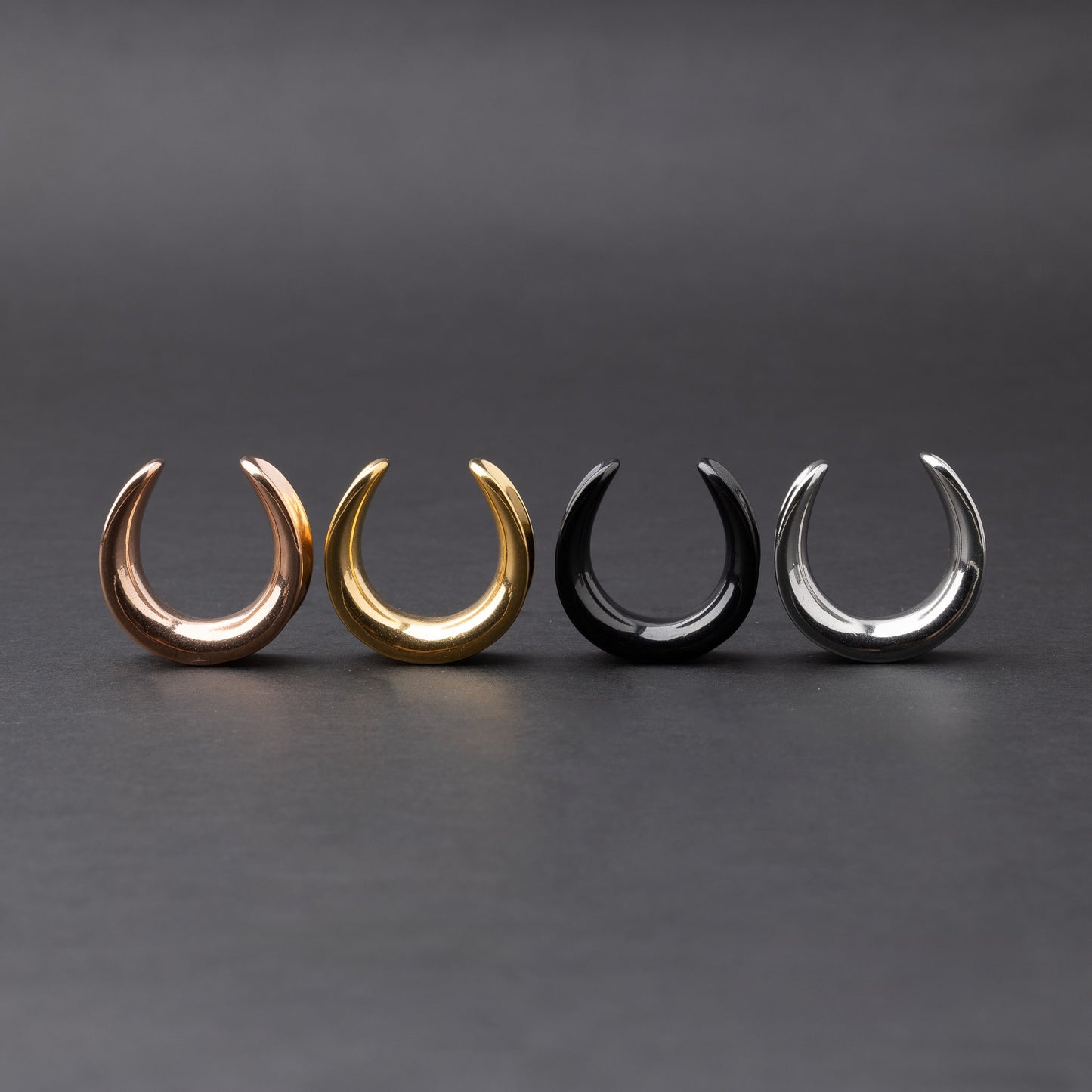 Polished saddle-style tunnels in rose gold, gold, black, and silver finishes, arranged on a matte dark background, showcasing their sleek and minimalist design.