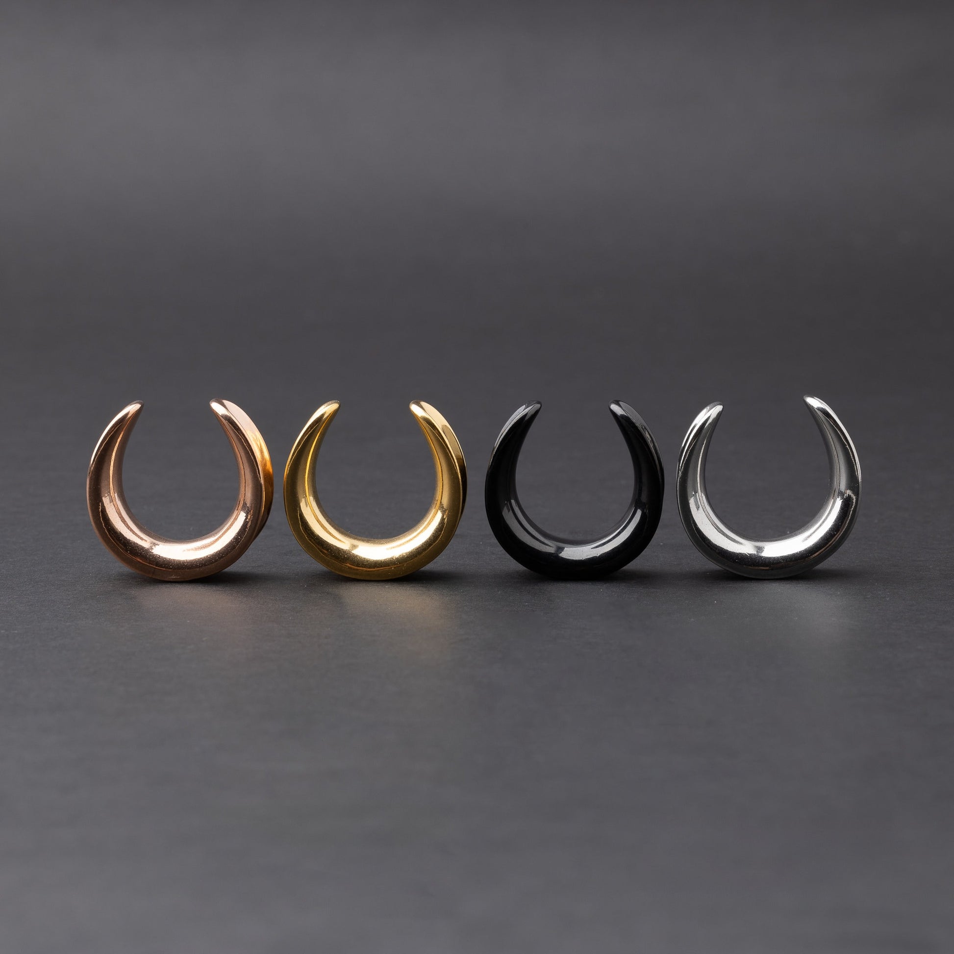 Polished saddle-style tunnels in rose gold, gold, black, and silver finishes, arranged on a matte dark background, showcasing their sleek and minimalist design.