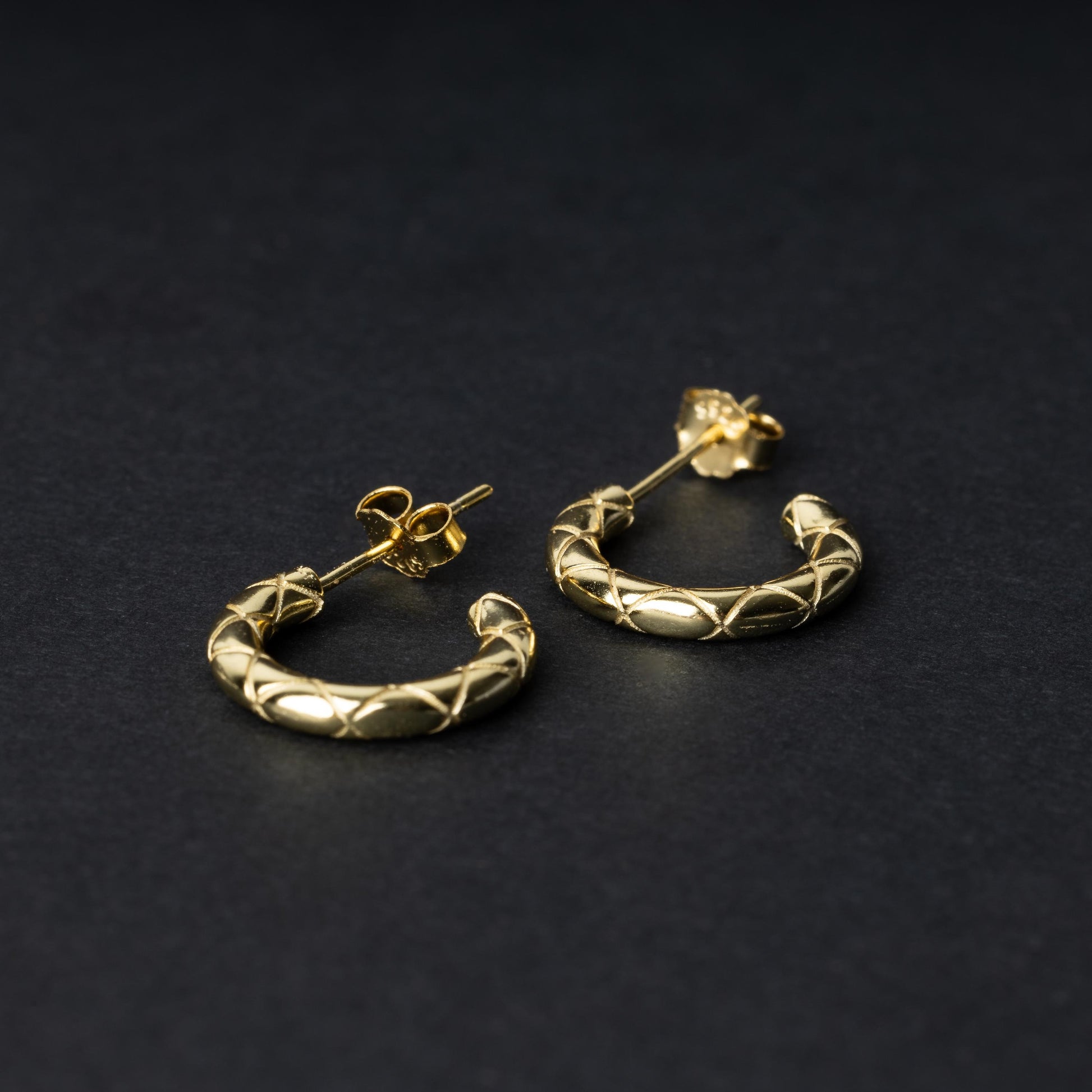 A pair of small gold quilted hoop earrings with post backs displayed on a dark background.
