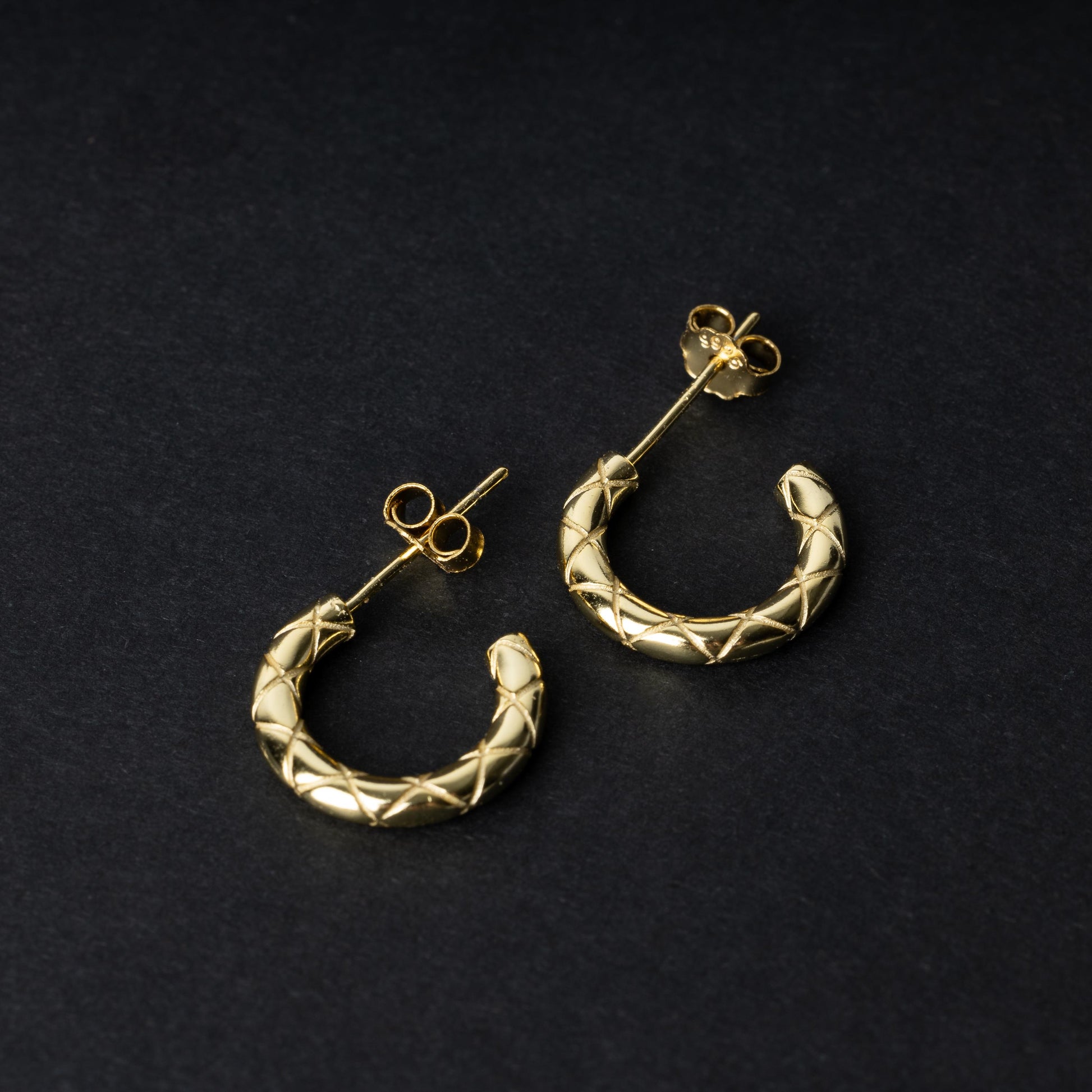 A pair of small gold quilted hoop earrings with post backs displayed on a dark background.