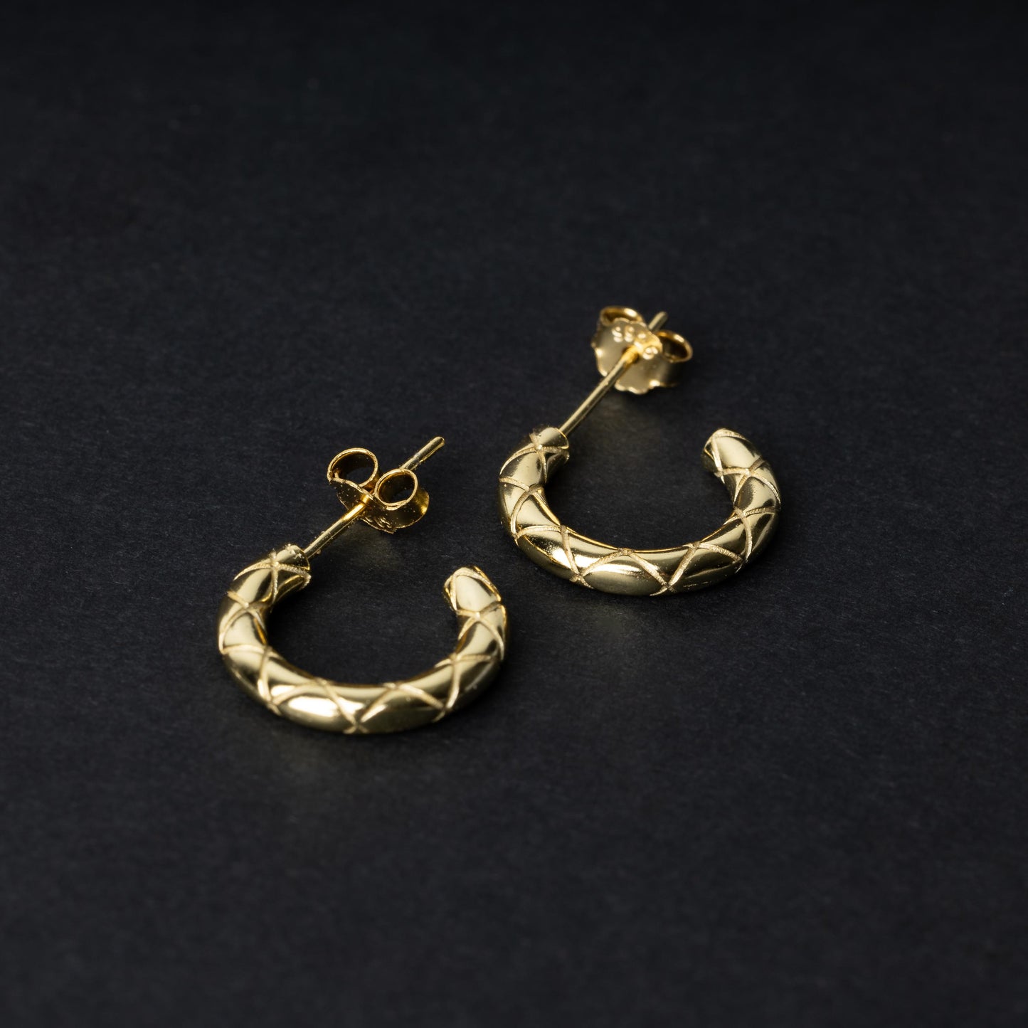 A pair of small gold quilted hoop earrings with post backs displayed on a dark background.
