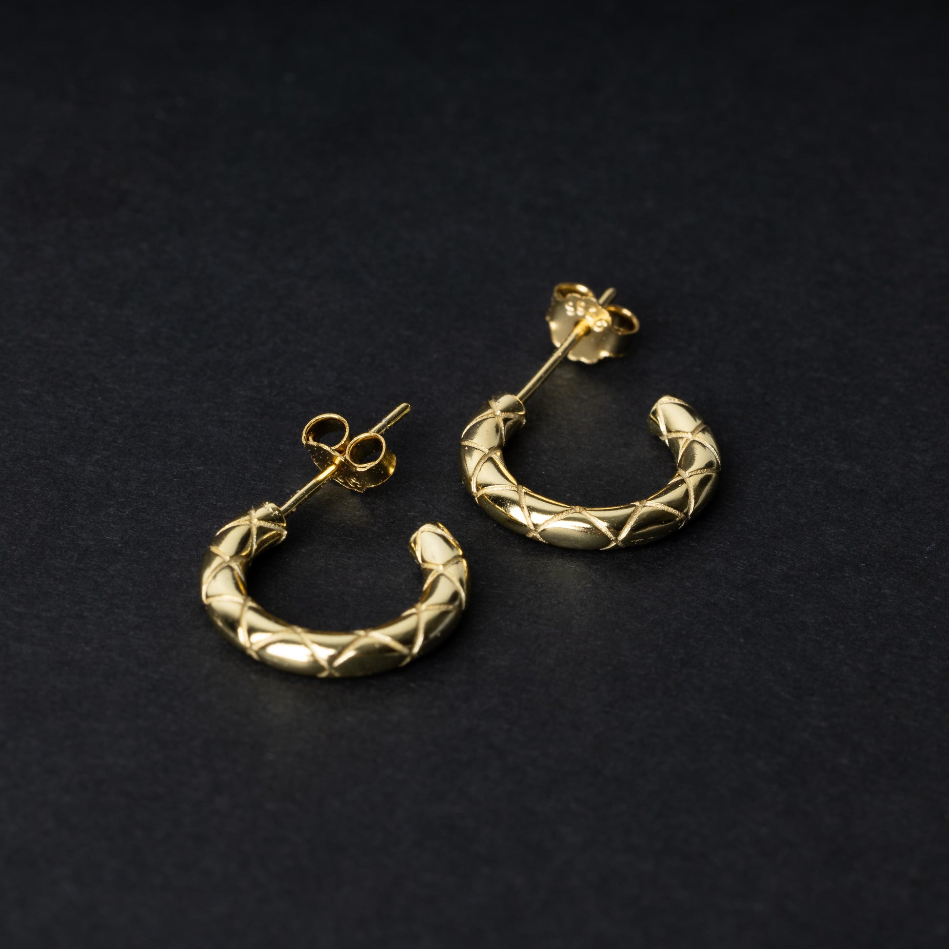 A pair of small gold quilted hoop earrings with post backs displayed on a dark background.