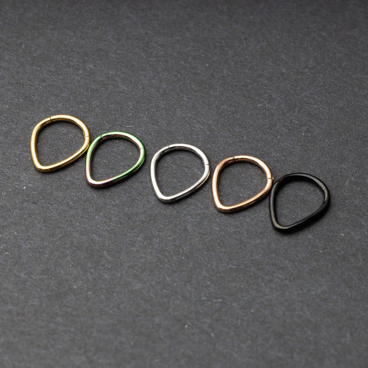 A collection of teardrop-shaped clickers in gold, rainbow, silver, rose gold, and black finishes arranged on a dark background.