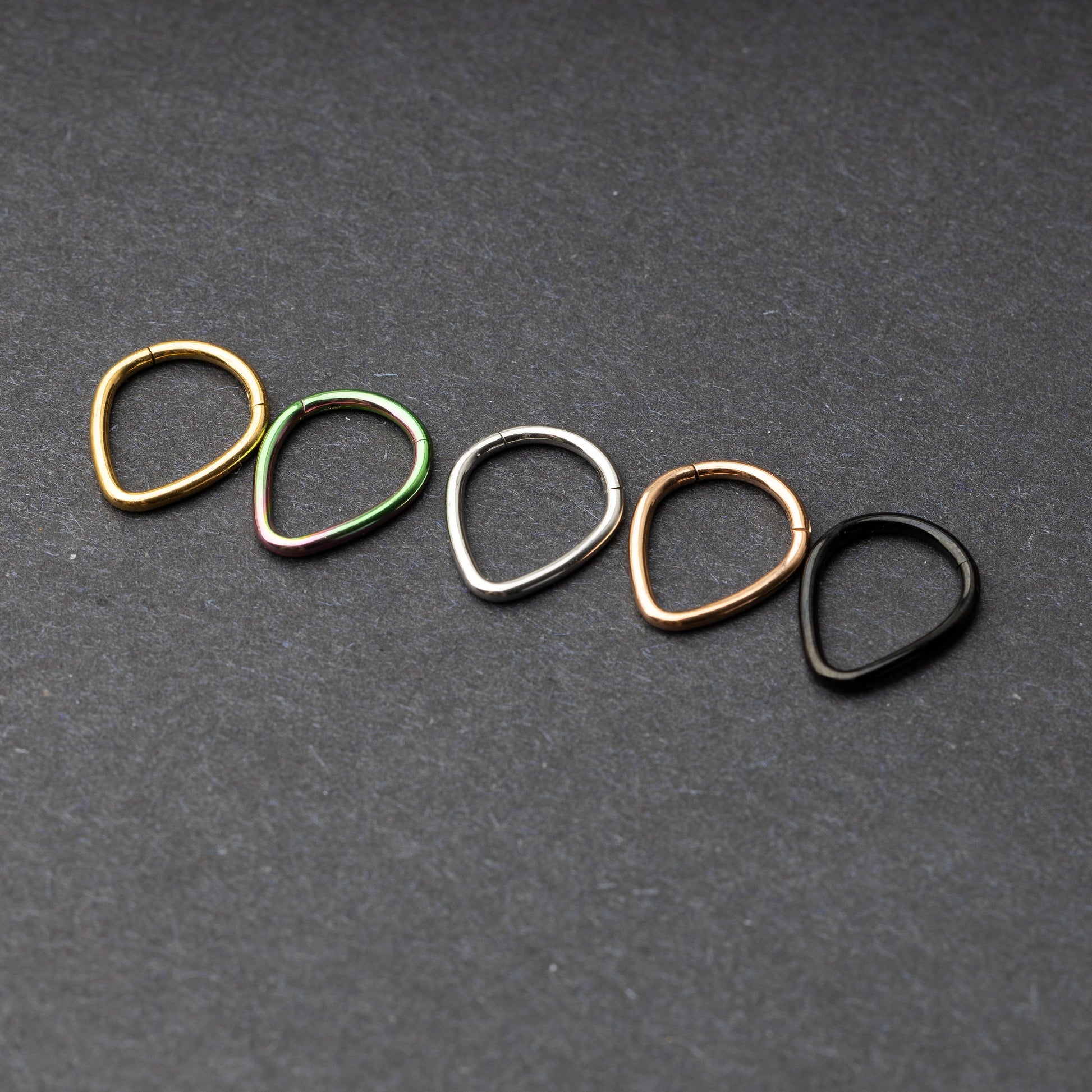 A collection of teardrop-shaped clickers in gold, rainbow, silver, rose gold, and black finishes arranged on a dark background.