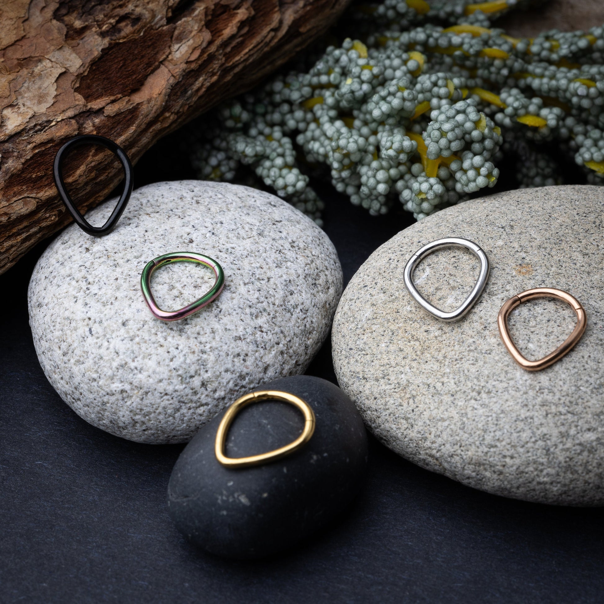 A collection of teardrop-shaped clickers in gold, rainbow, silver, rose gold, and black finishes arranged on a stone and wooden background.