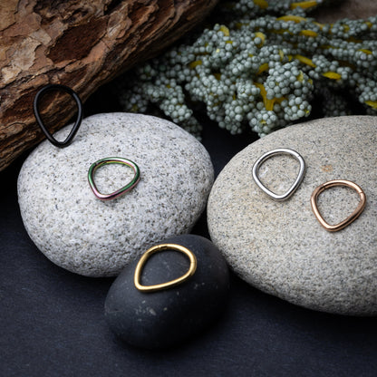 A collection of teardrop-shaped clickers in gold, rainbow, silver, rose gold, and black finishes arranged on a stone and wooden background.