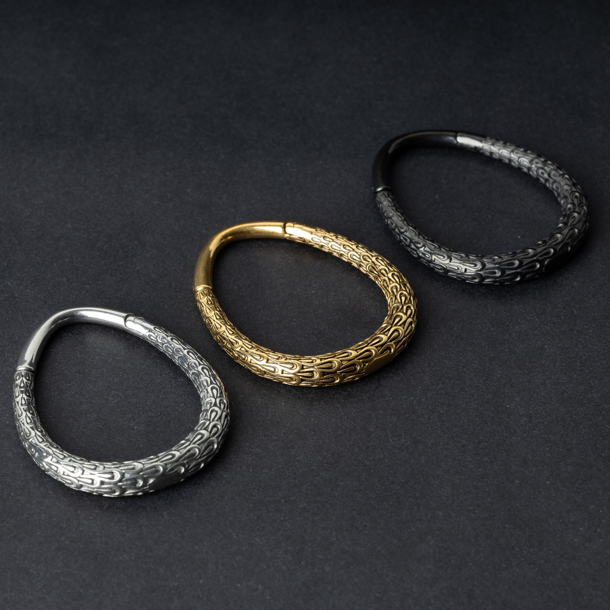 Three teardrop-shaped ear weights with a pebble-textured finish in silver, gold, and black, displayed on a dark background.

