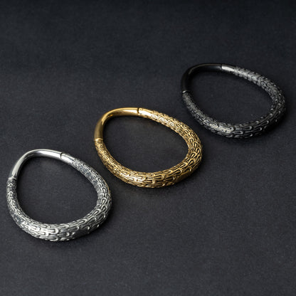 Three teardrop-shaped ear weights with a pebble-textured finish in silver, gold, and black, displayed on a dark background.

