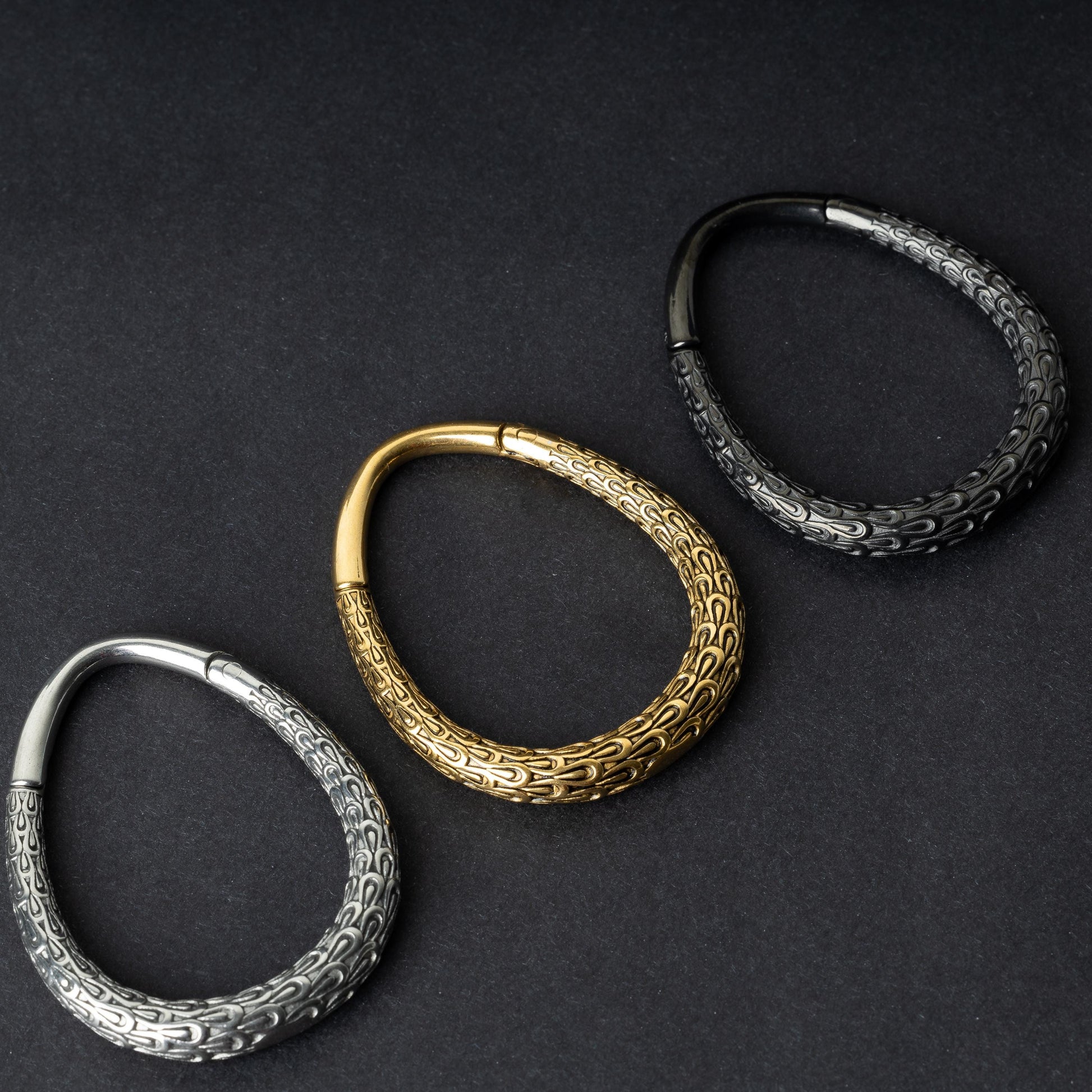 Three teardrop-shaped ear weights with a pebble-textured finish in silver, gold, and black, displayed on a dark background.

