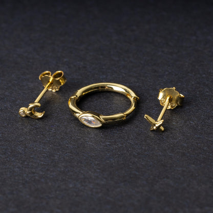 A gold-tone earring set displayed on a dark matte background, featuring a hoop with a marquise-cut crystal, a crescent moon stud with a crystal accent, and a minimalist cross stud.
