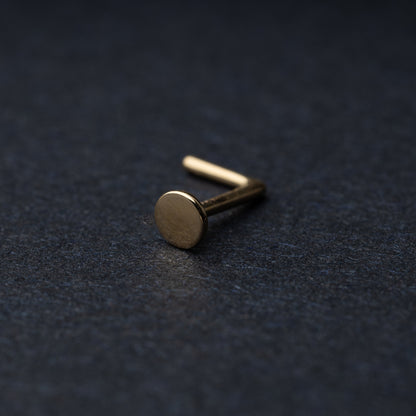 Solid gold nose stud with a flat disc design, displayed on a dark textured background.