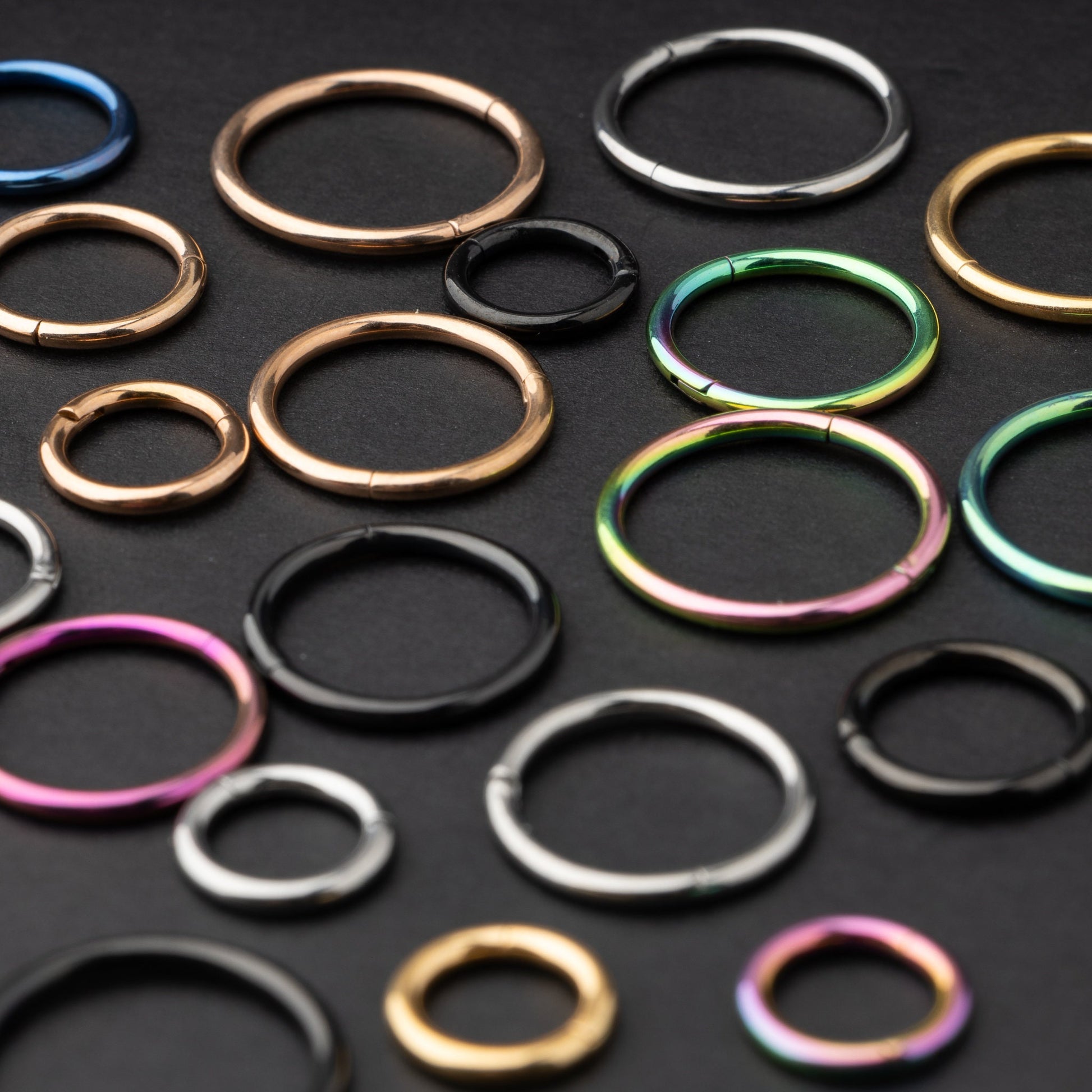 Assorted titanium seam rings in a variety of colors, including gold, black, silver, and rainbow, displayed on a dark textured background.

