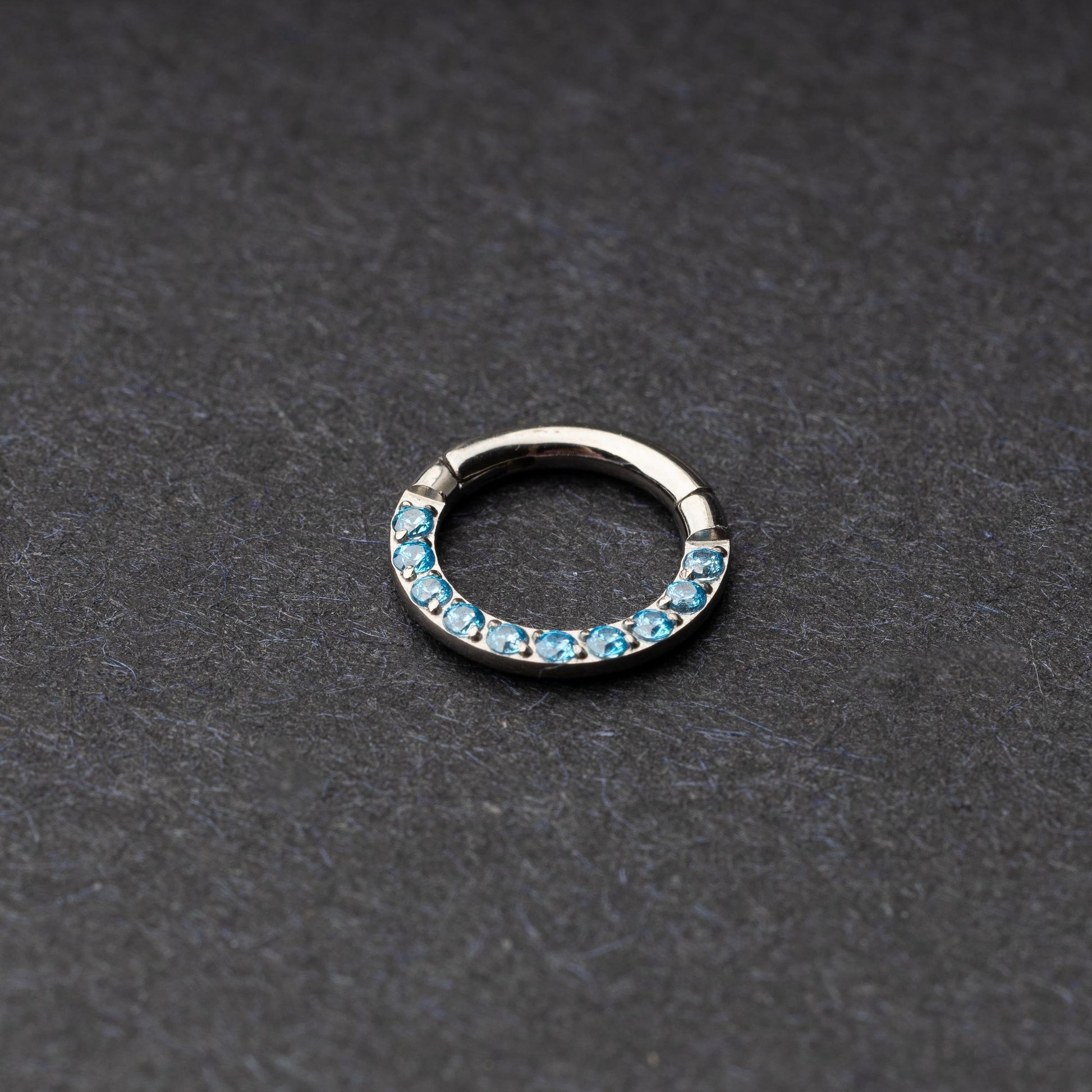 Stainless steel septum clicker with bright blue gemstones, displayed on a dark textured background.