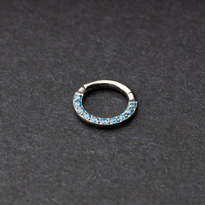 Stainless steel septum clicker with bright blue gemstones, displayed on a dark textured background.
