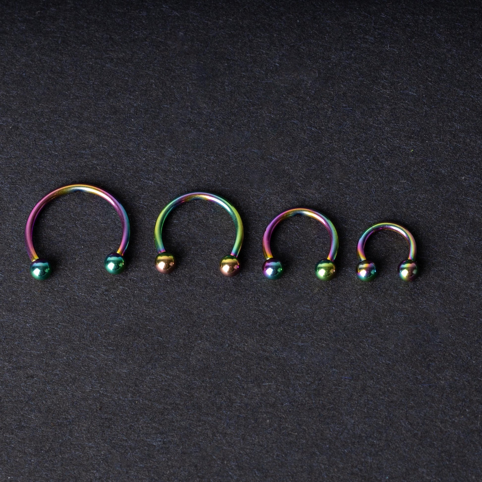 Close-up of four anodized rainbow horseshoe rings in varying sizes, with vibrant, multicolor finishes, displayed on a dark background.

