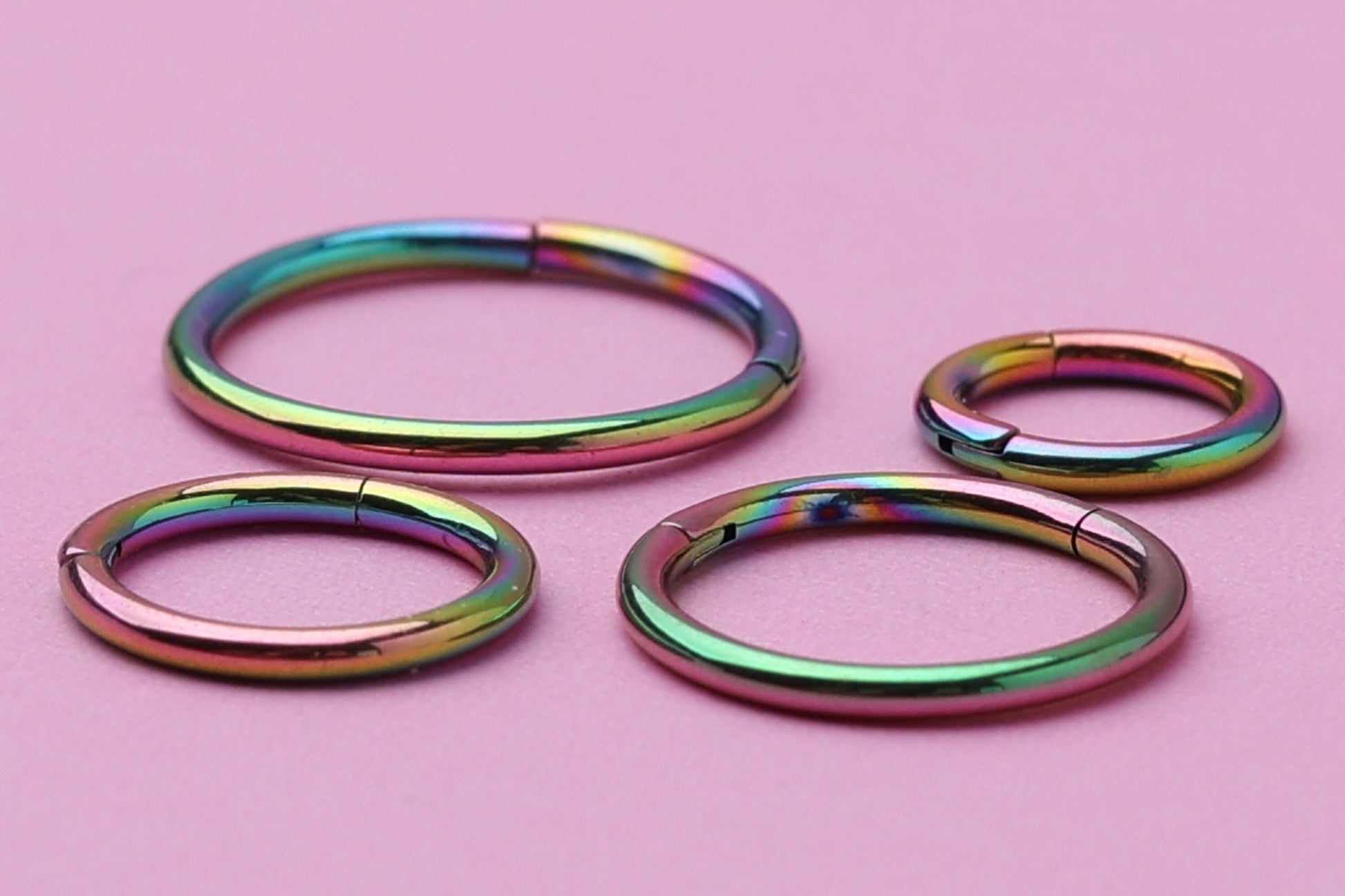  A collection of titanium seamless hoops in a vibrant rainbow finish, displayed on a Pink textured background in varying sizes.