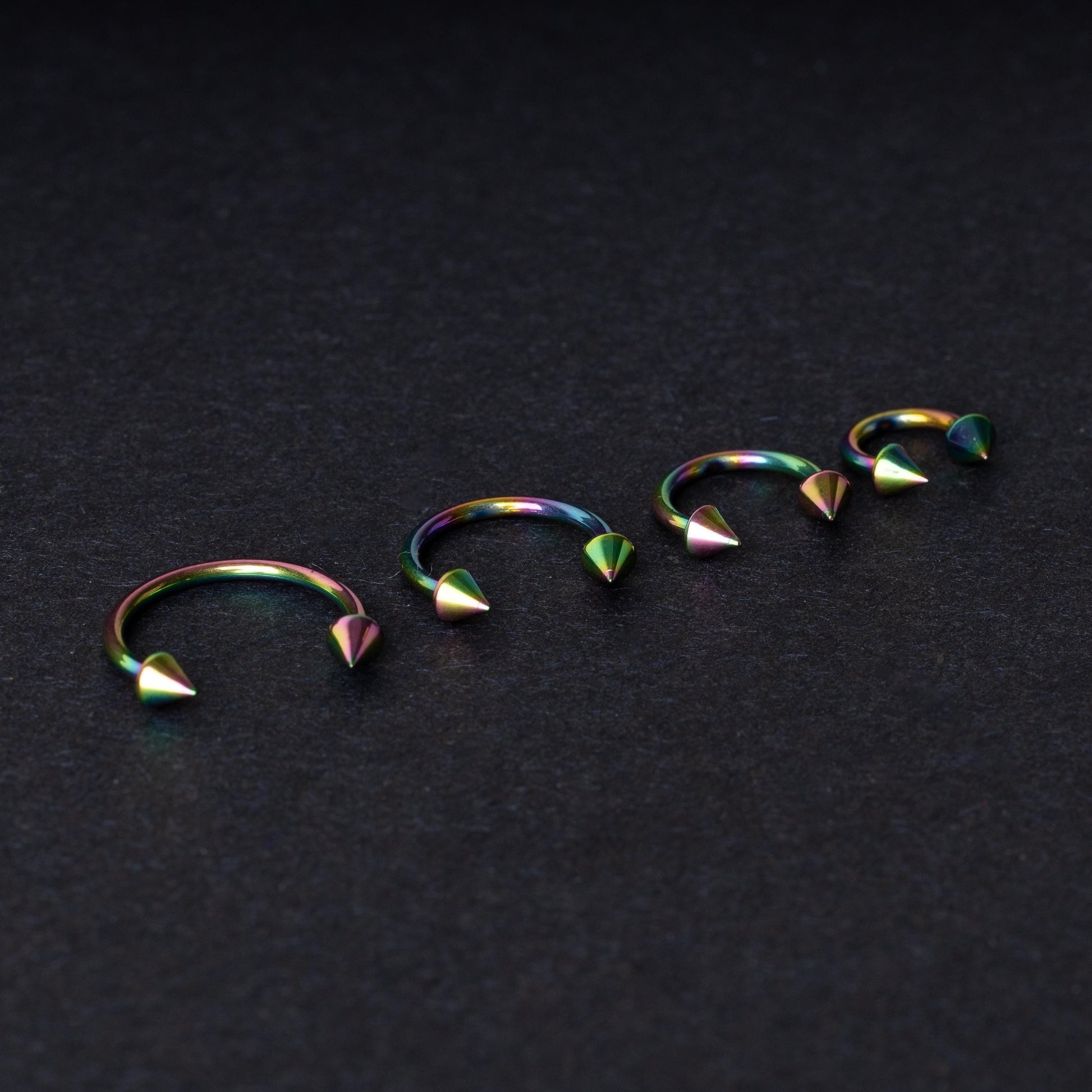 Titanium horseshoe hoops with spike ends, featuring a colorful rainbow finish, displayed on a dark textured background.