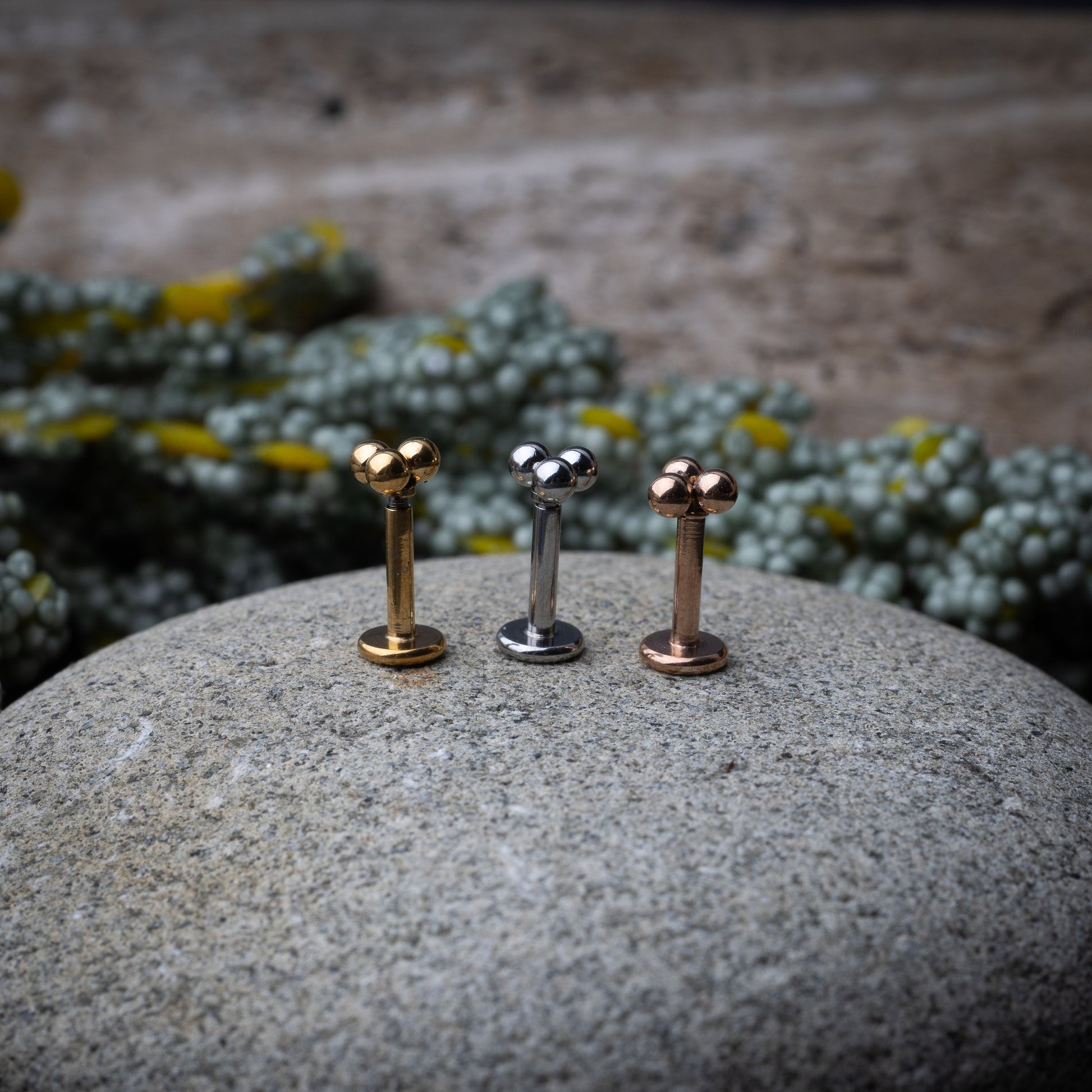 Close-up of three minimalist ball cluster labret studs in rose gold, silver, and gold finishes, displayed on a stone background.

